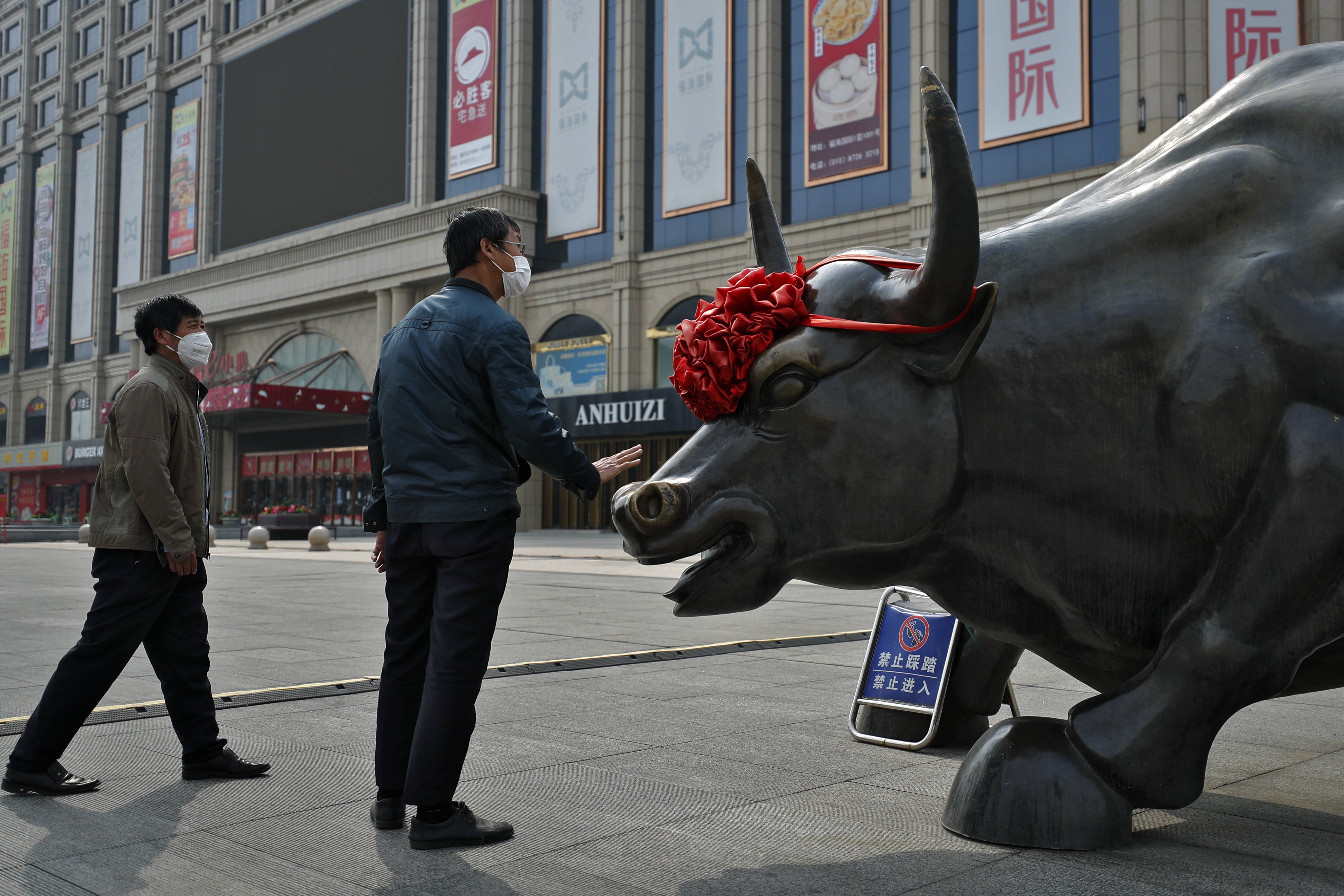 A man wearing a protective face mask touches the investment icon bull statue on display outside a mall in Beijing in March 2020. Photo: AP