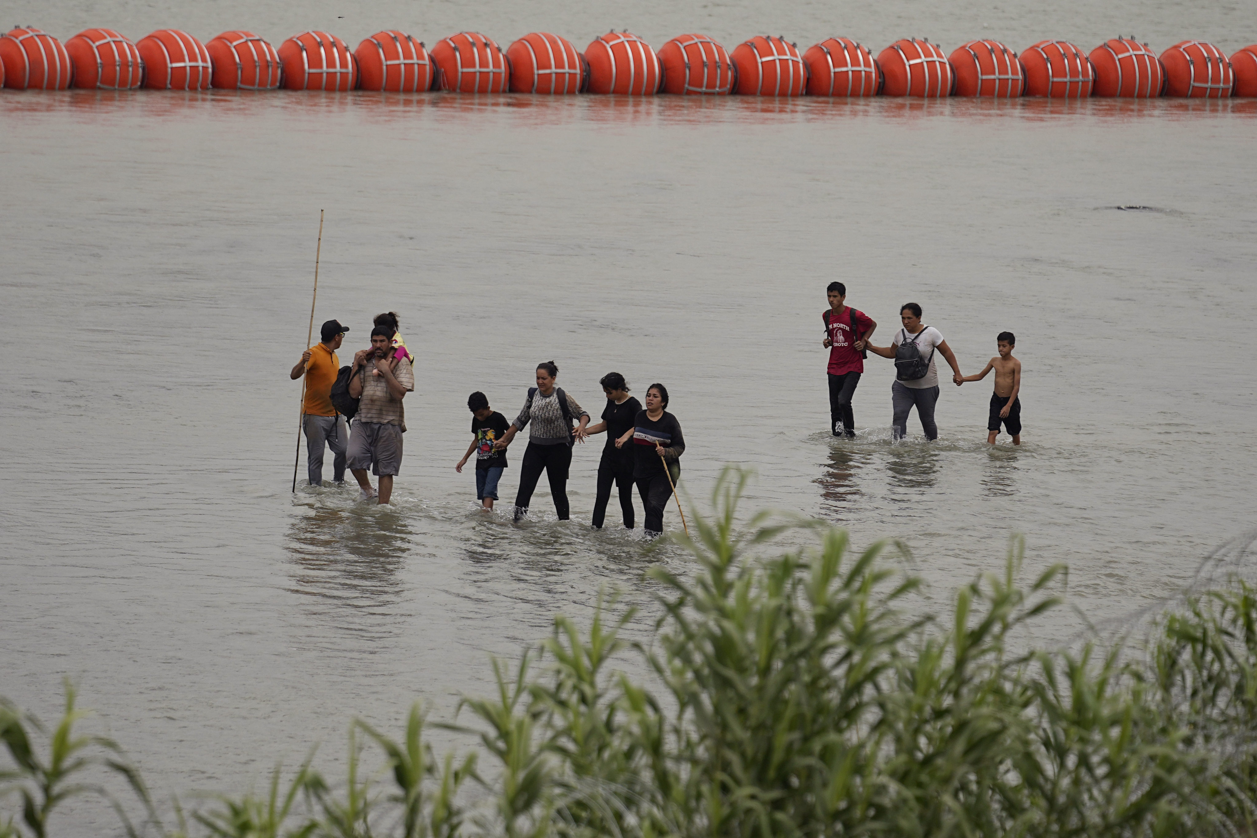 Migrants who crossed the Rio Grande from Mexico walk past large buoys being deployed as a border barrier on the river in Eagle Pass, Texas. Photo: AP