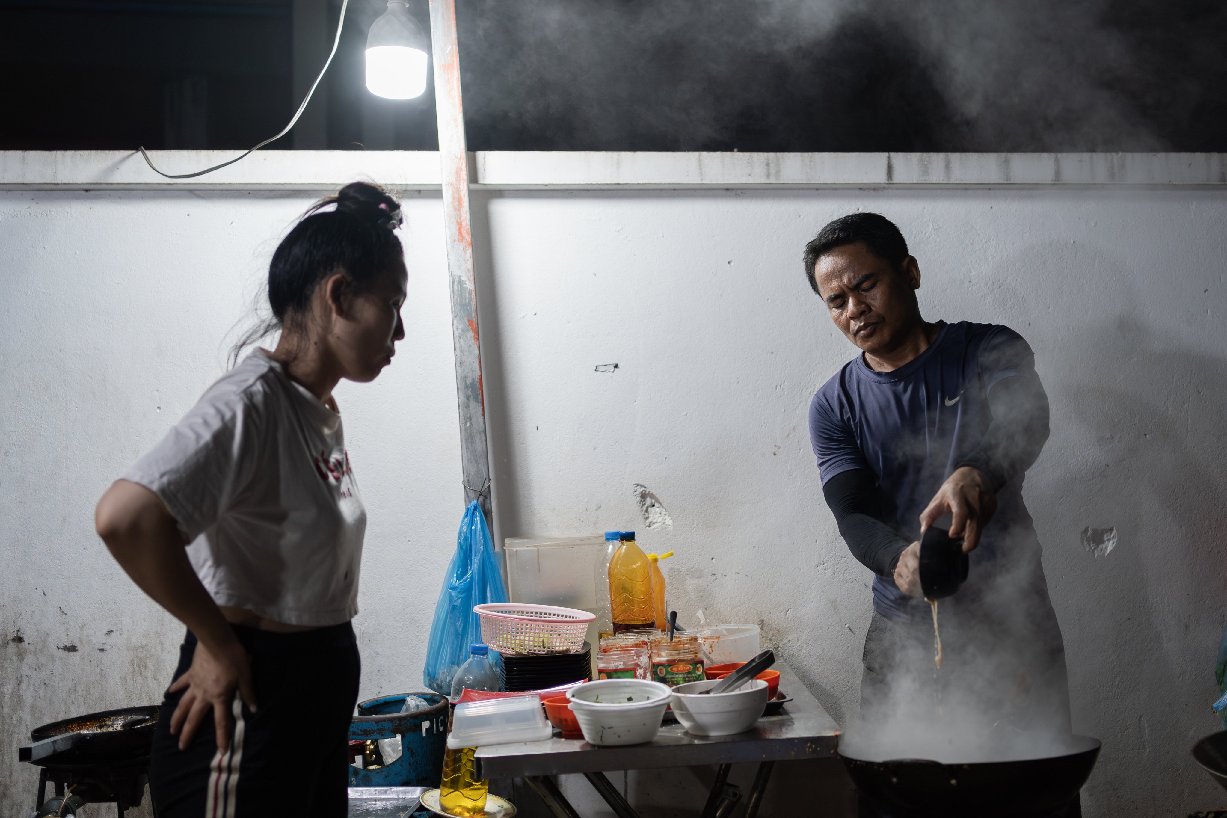 18th June, 2023 - Siem Reap (Cambodia). Kalida watches as her husband Chin Meas fries noodles for their customers. In 2015, Meas moved to Siem Reap to set up a noodle store with his wife. Now, cooking noodles 14 hours a day every day to make ends meet, the renowned poet struggles to find time to write poetry anymore. Credit: Andy Ball