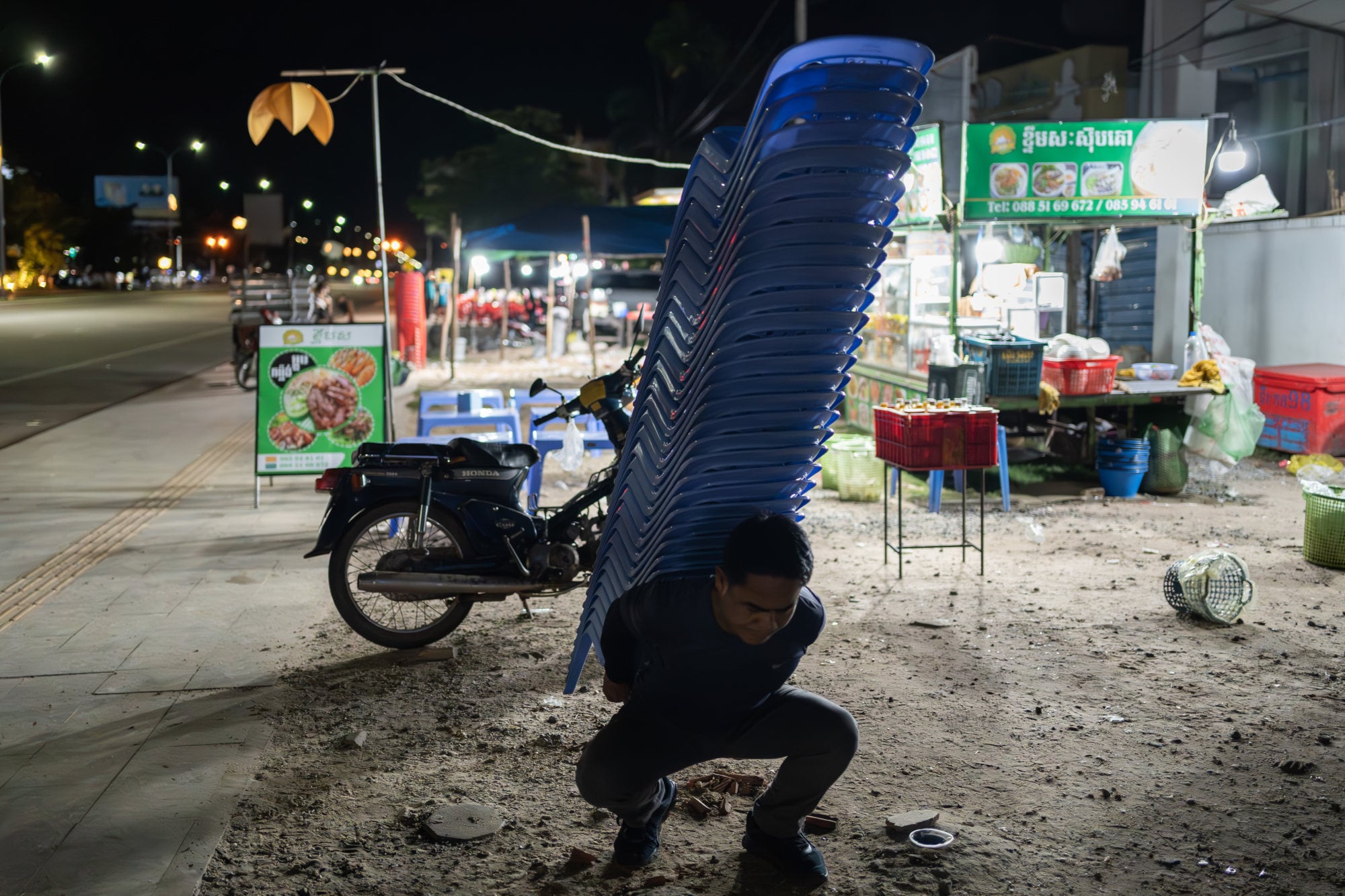 Meas stacks plastics chairs on his back as he packs away the noodle stand. Photo: Andy Ball