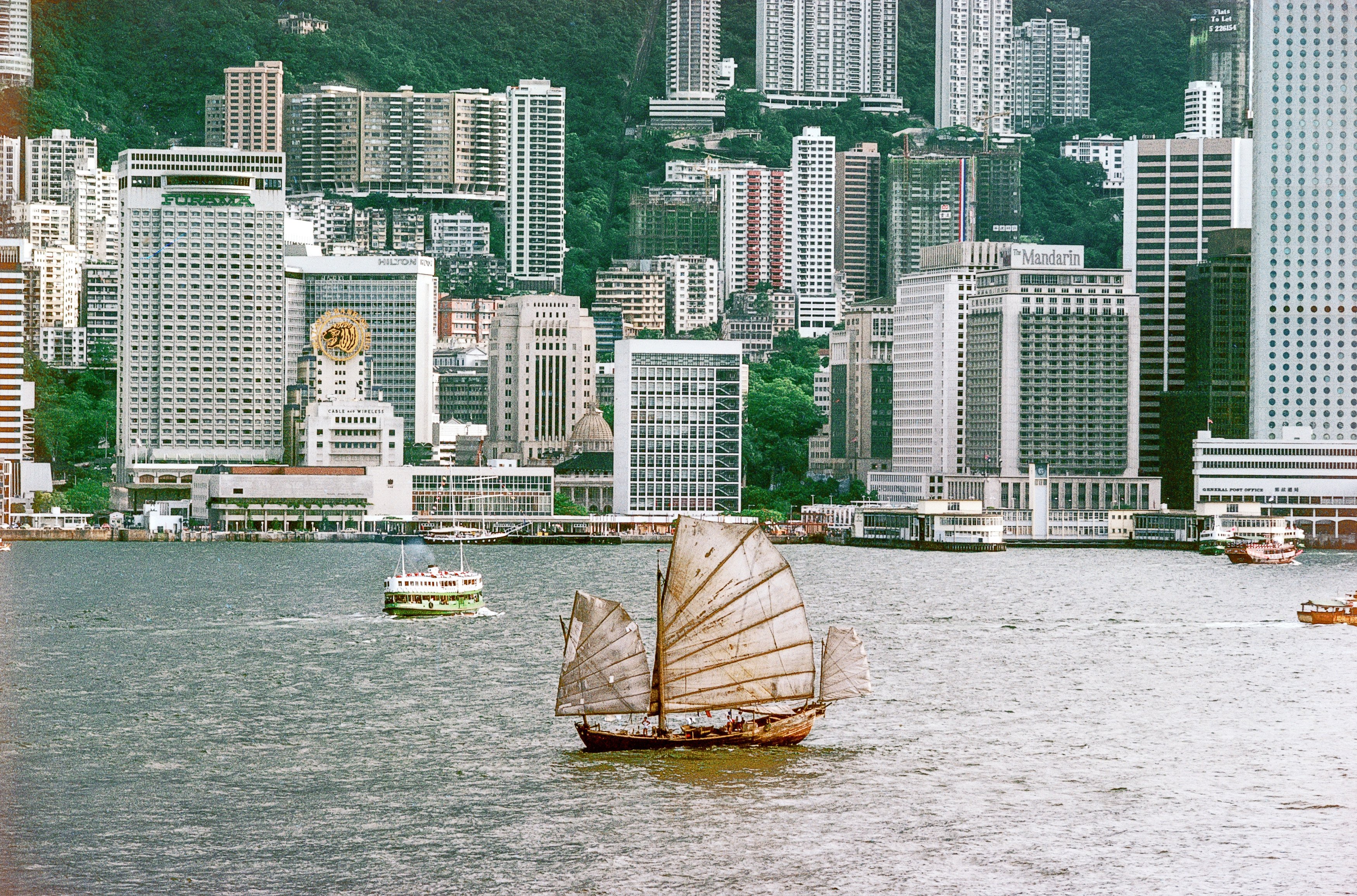 A lone junk plies Victoria Harbour in 1982,  by which time the fleets of junks that once plied its waters were a thing of the past. The Hong Kong Tourism Board adopted a junk as a symbol of the city with great success, but it long ago ceased representing reality.