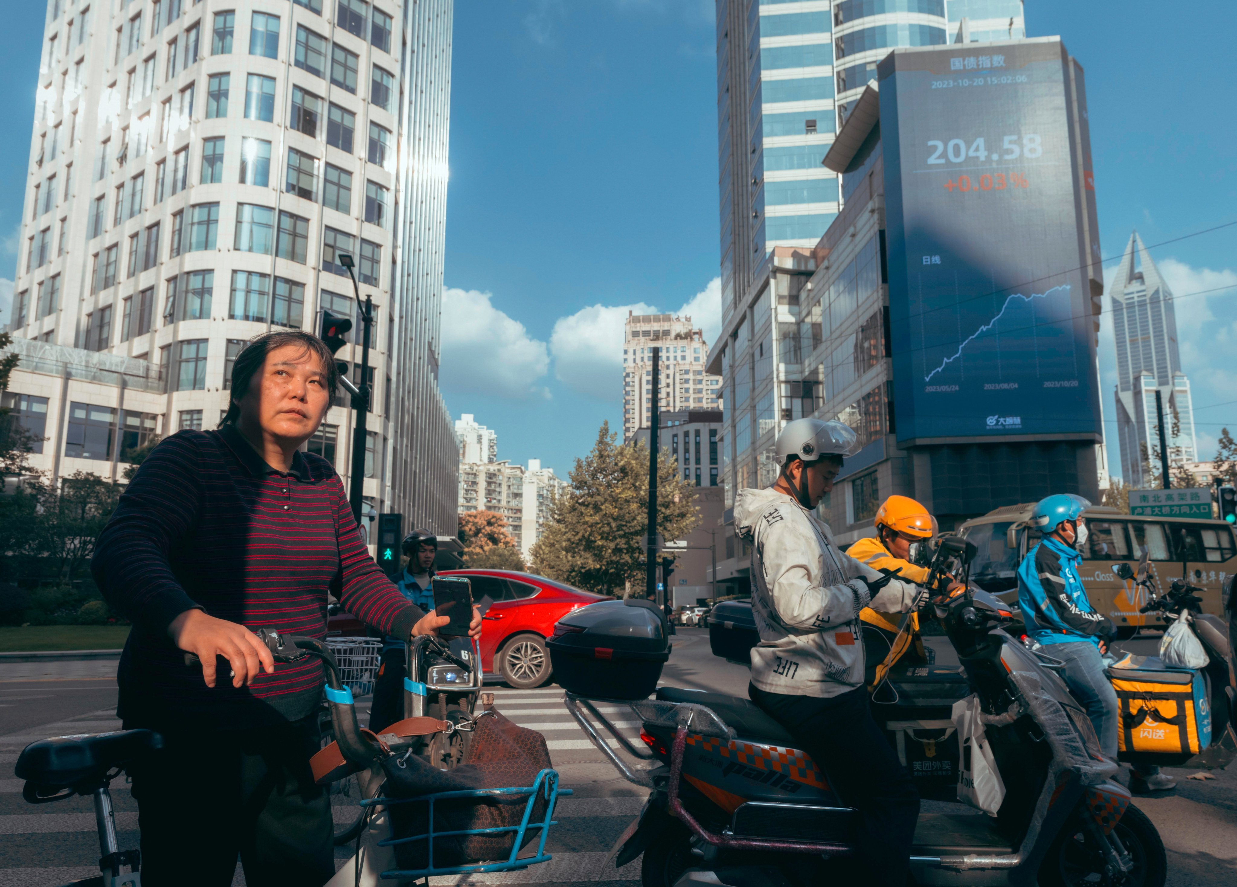 People riding bicycles and scooters on the street beneath a screen showing the latest stock and economy data in Shanghai in October 2023. Photo: EPA-EFE