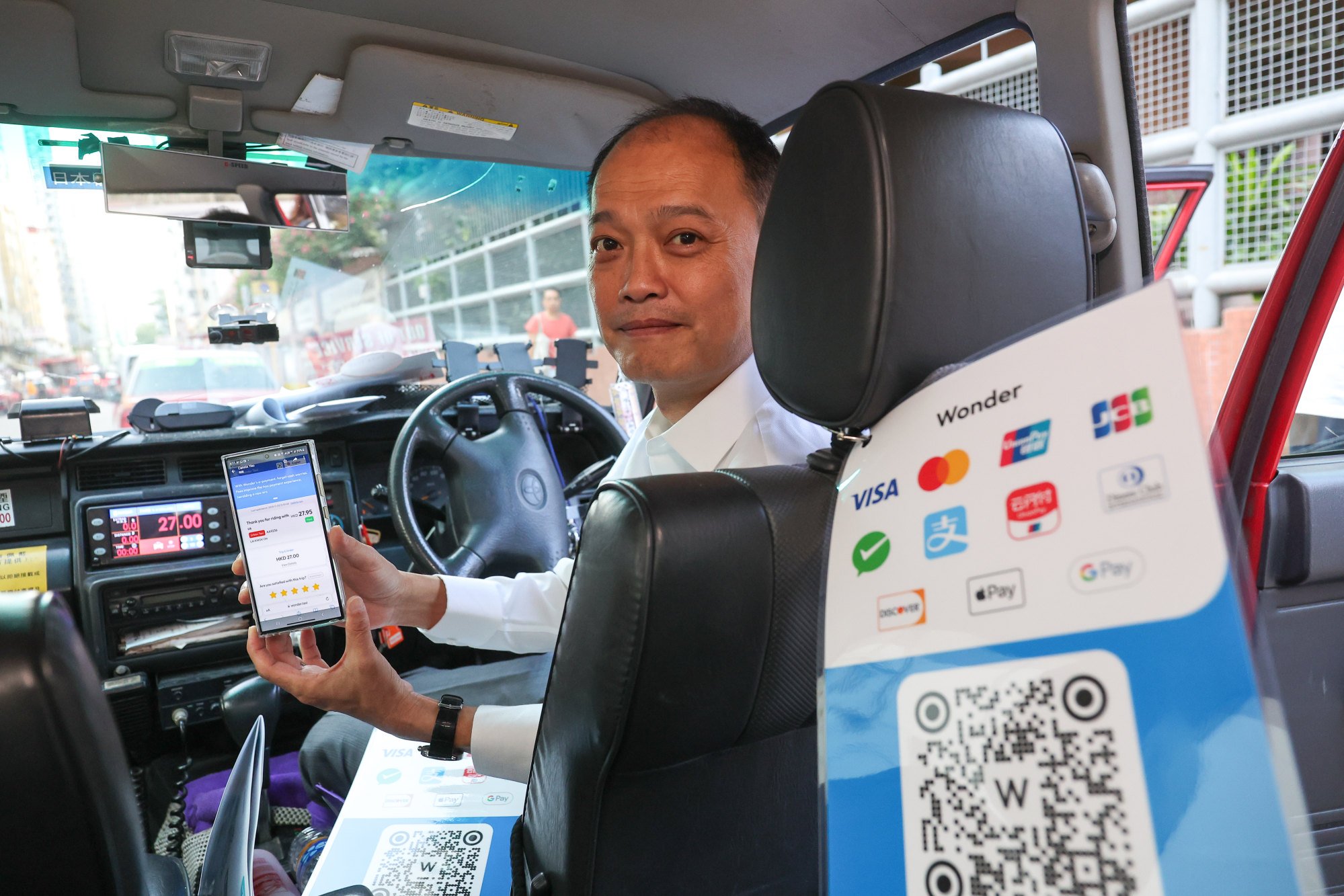A Hong Kong taxi driver is sitting in his vehicle, holding a mobile device displaying a payment confirmation, with a dashboard-mounted device and a sticker showing various payment options and QR codes visible in the foreground.