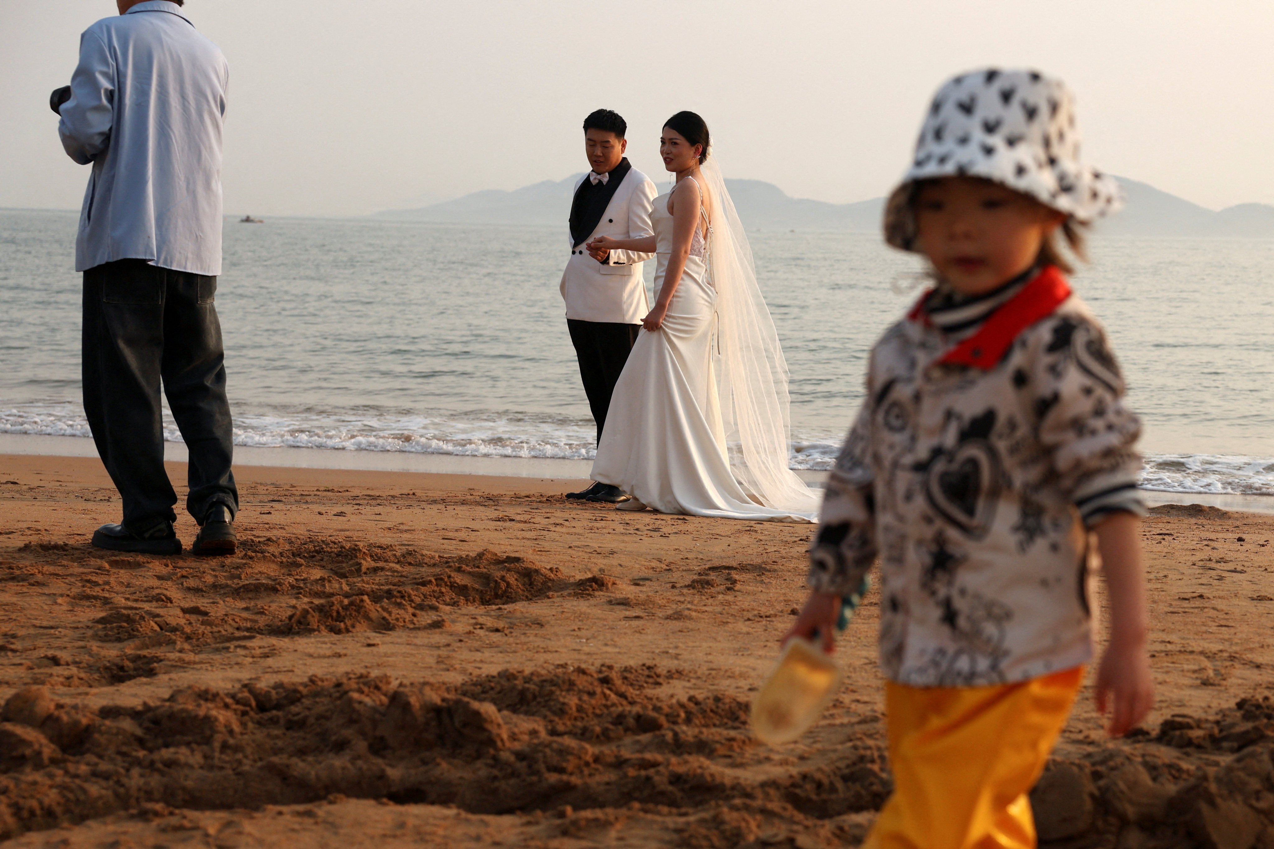 A child plays with sand near a couple taking part in a pre-wedding photoshoot on a beach in Qingdao, Shandong province. Photo: Reuters