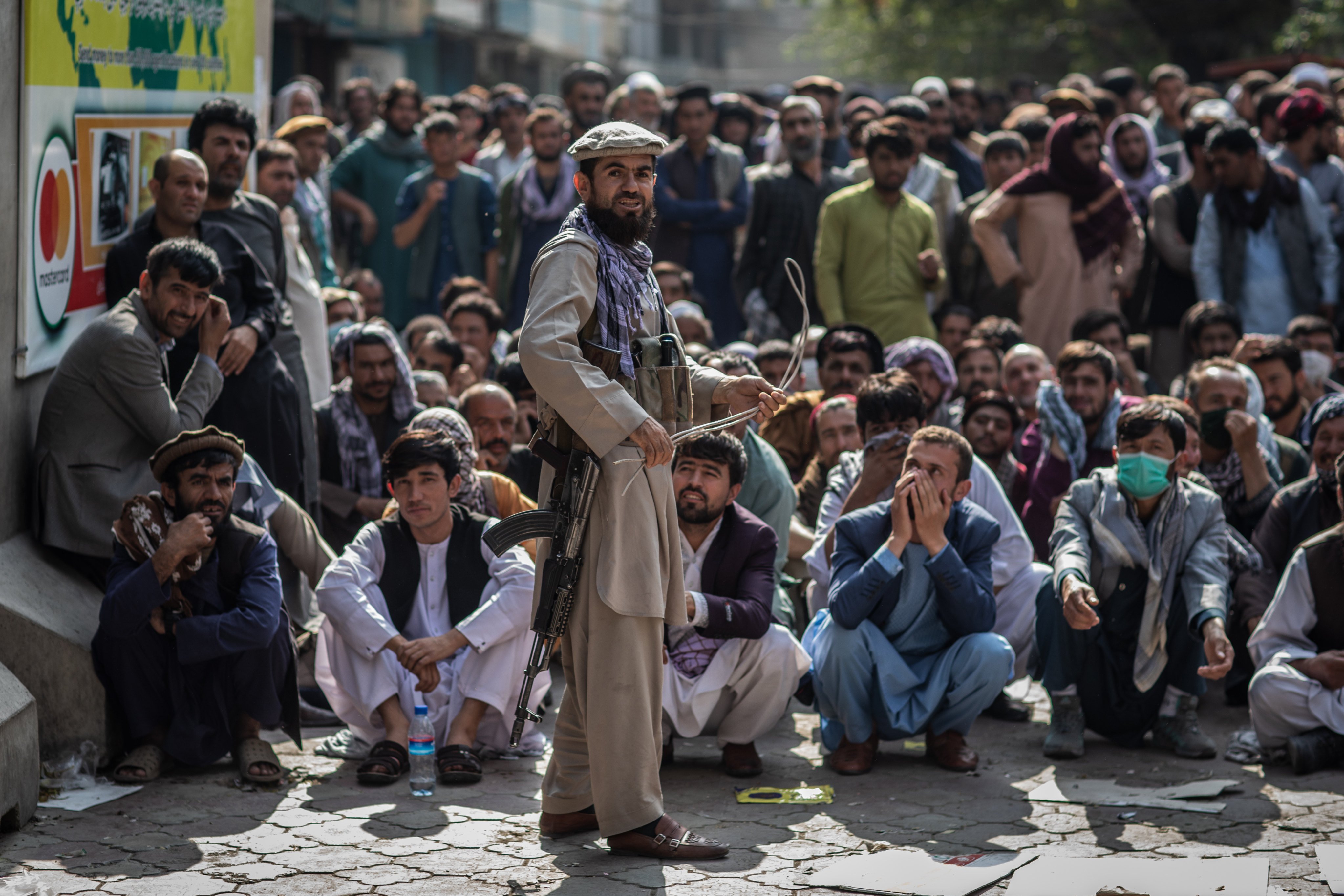 A Taliban security guard stands guard with with a whip in front of Afghan men waiting outside a bank to withdraw money. Photo: dpa