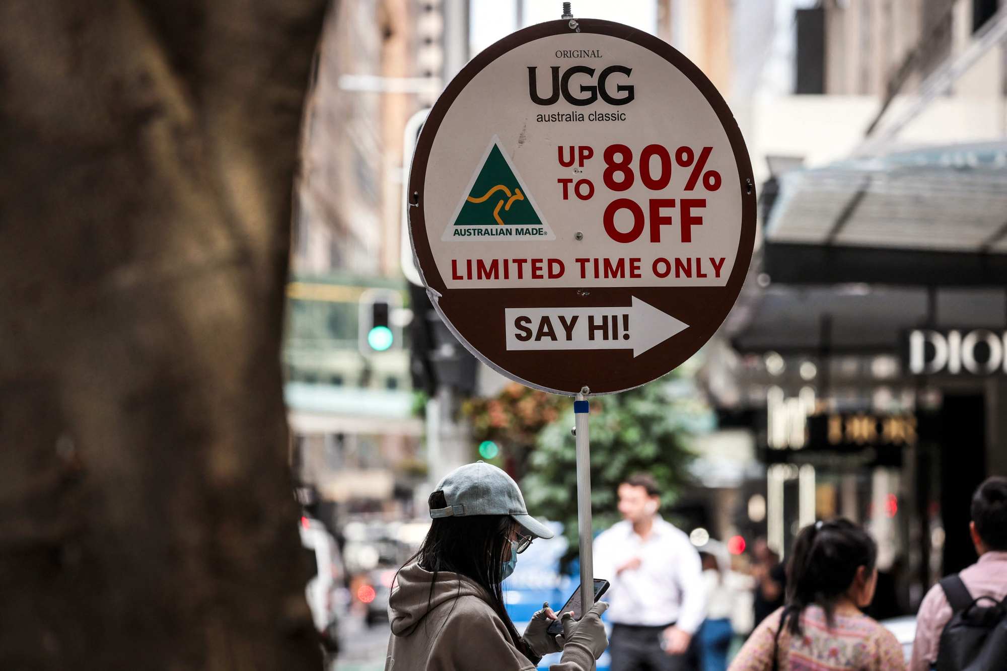 A woman looks at her phone as she holds a sign advertising Australian-made UGG boots for sale in central Sydney on April 10. Australia has been hit with a blanket 10 per cent tariff on goods exported to the United States. Photo: AFP A woman looks at her phone as she holds a sign advertising Australian-made UGG boots for sale in central Sydney on April 10. Australia has been hit with a blanket 10 per cent tariff on goods exported to the United States. Photo: AFP