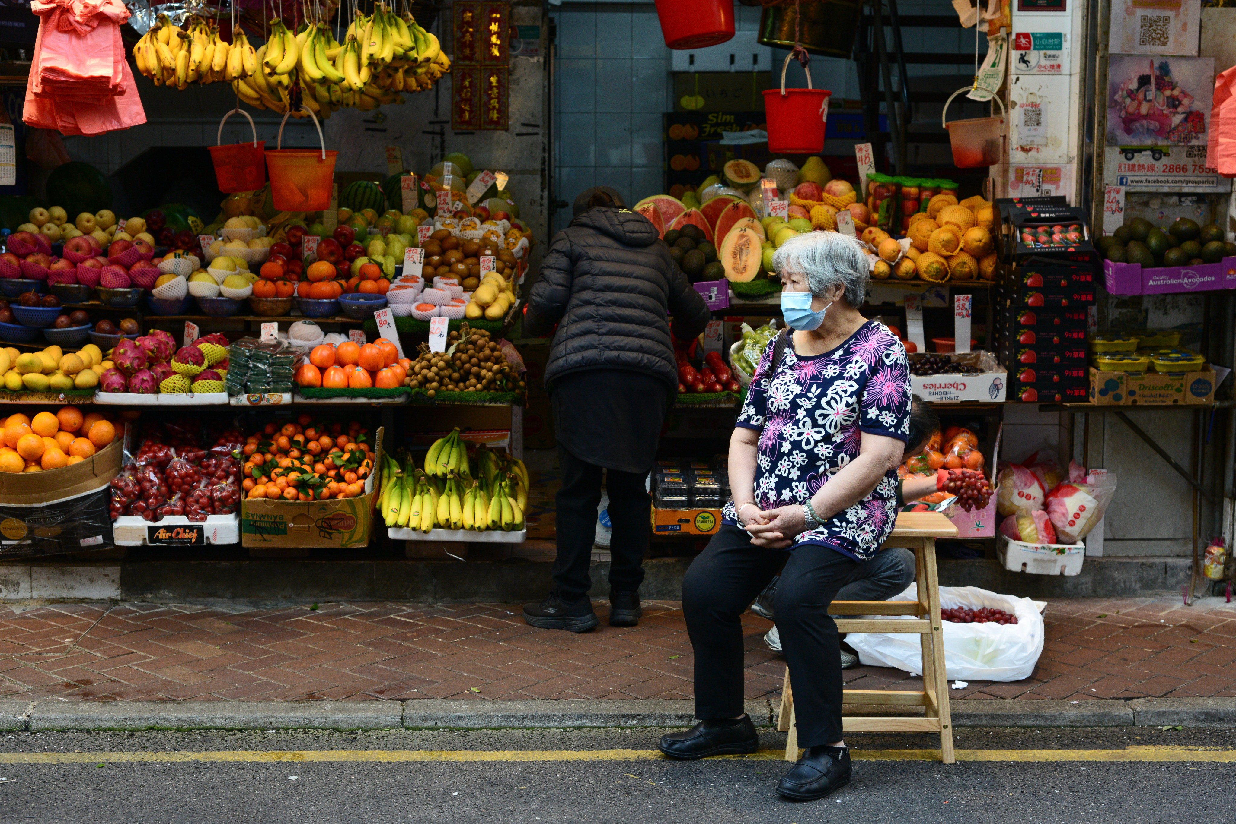 An elderly woman sits on a stool on the street in front of a fruit and vegetable seller in a market in Causeway Bay, Hong Kong, on December 26, 2024. Photo: Antony Dickson