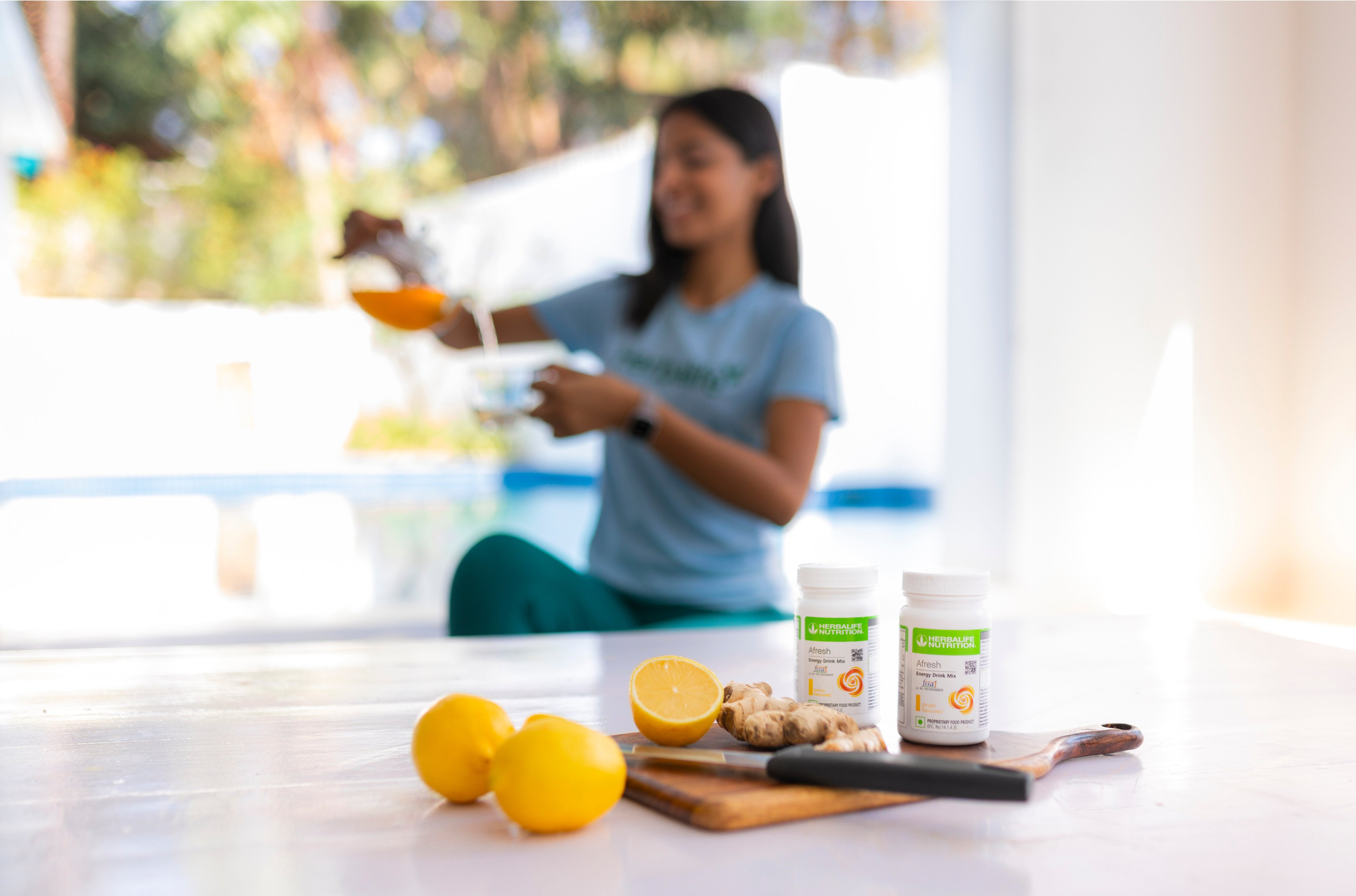 Woman smiling, pouring juice, with lemon, ginger, and supplements on a table in a bright kitchen.