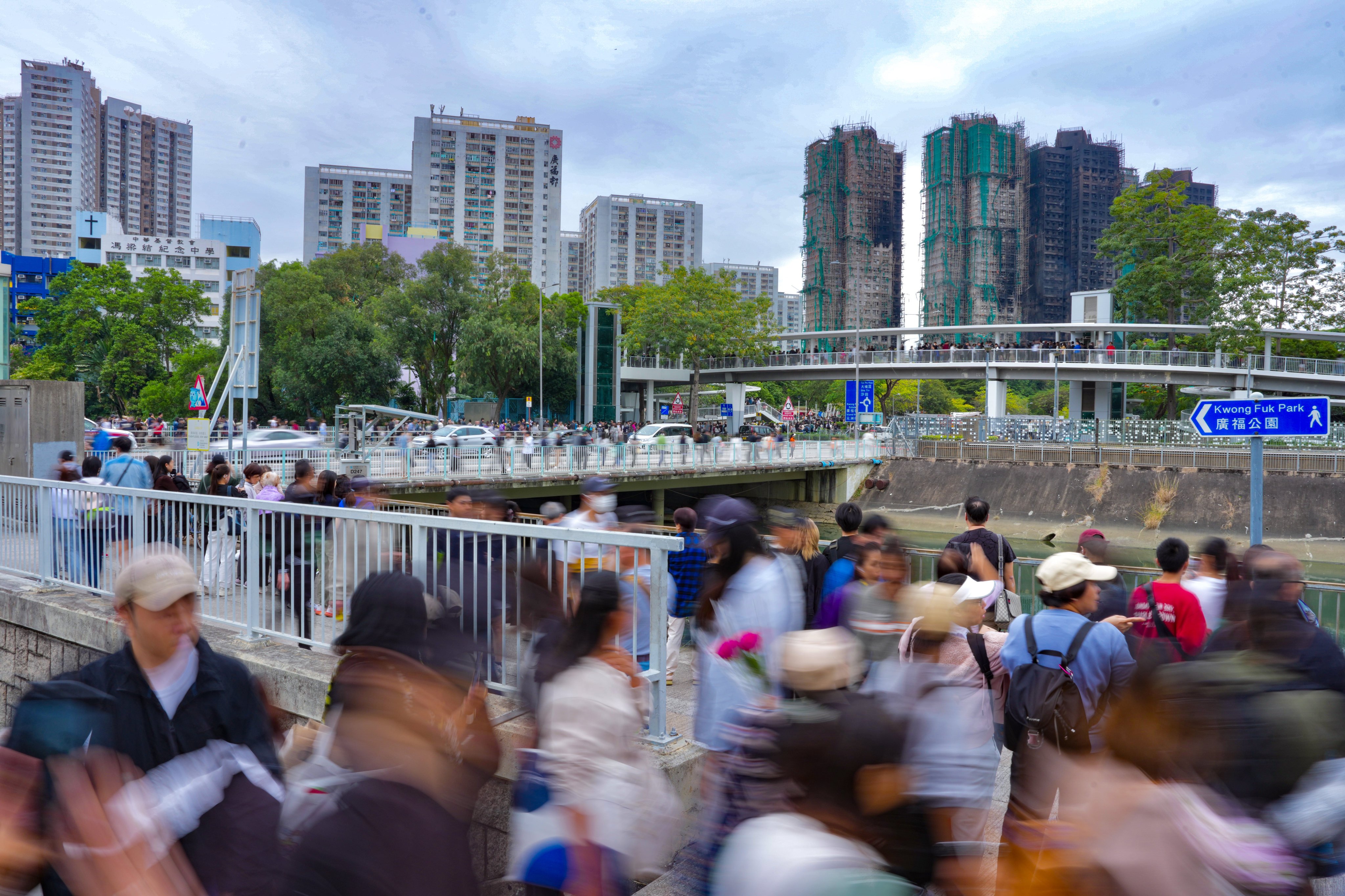 Hundreds of people queue on Sunday to lay flowers and pay tribute to victims of the deadly fire at Wang Fuk Court in Tai Po, seen in the background. Photo: Karma Lo
