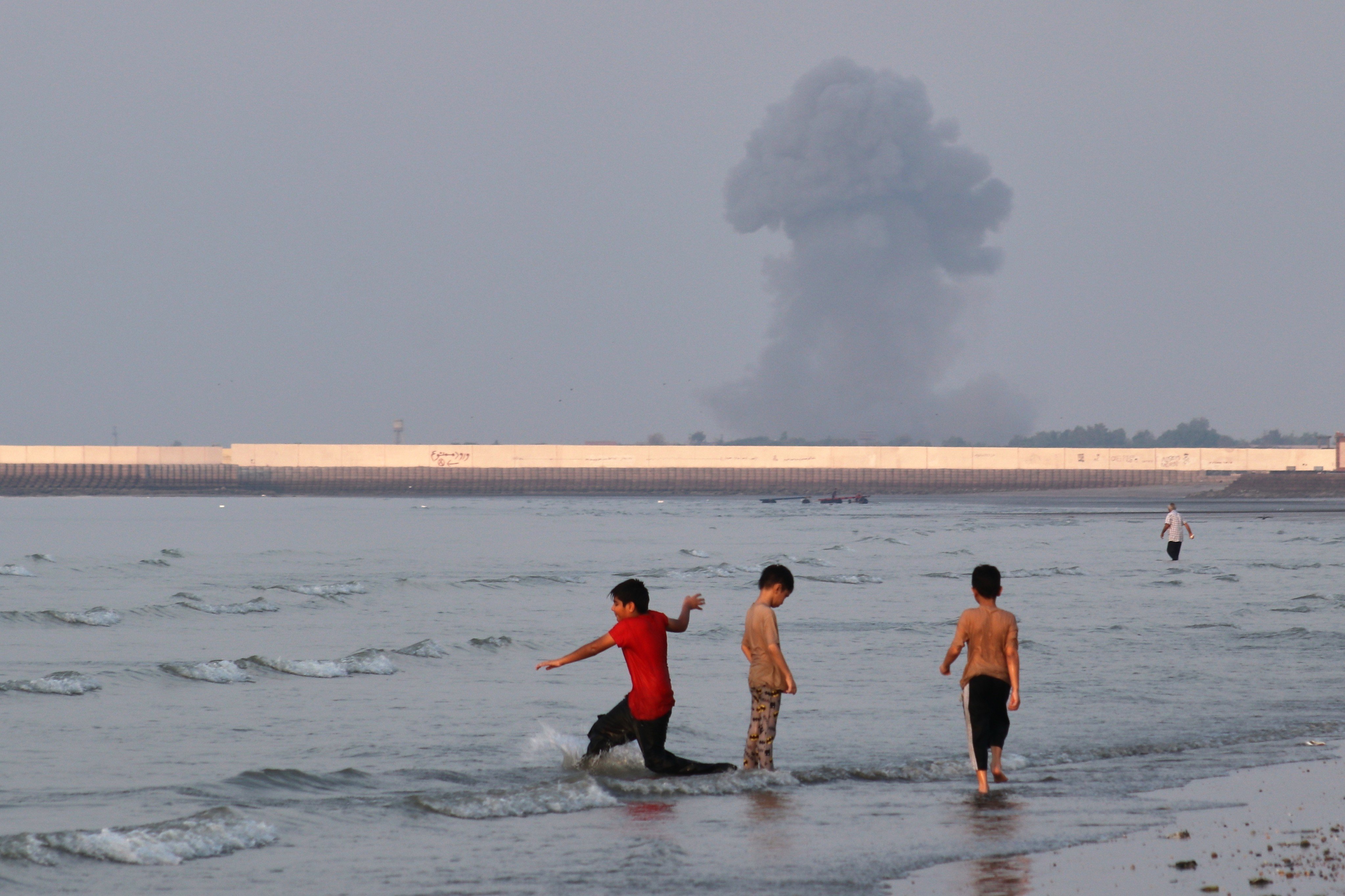 Boys play in the shallow waters of the Strait of Hormuz, as a plume of smoke rises from an explosion in the background, off Bandar Abbas, Iran, on Monday. Photo: ISNA via AP