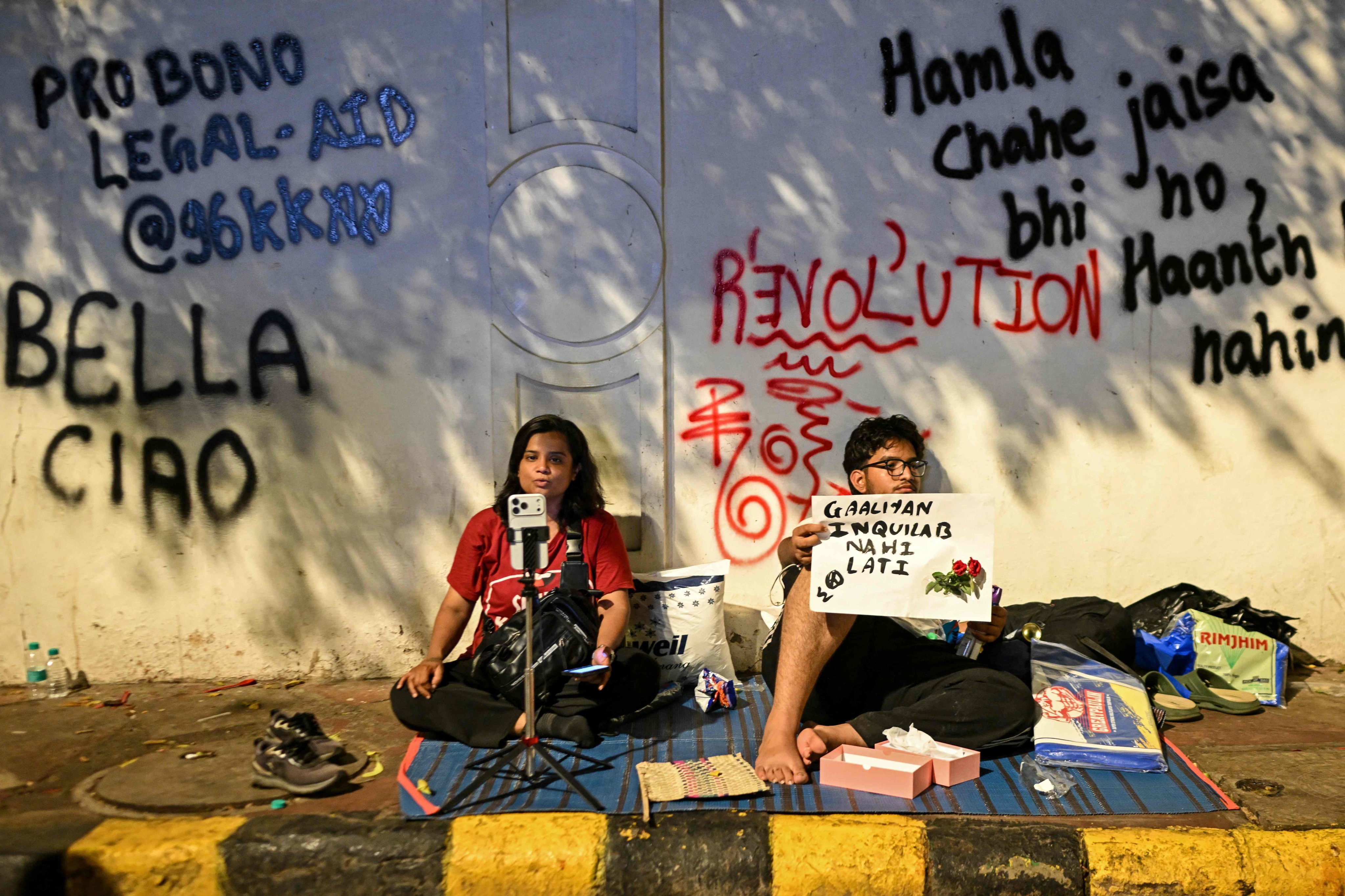 This photograph, taken on July 24, 2026, shows a Cockroach Janta Party (CJP) supporter recording a video while sitting beside graffiti against mainstream television media during a mass youth-led protest over exam irregularities at Jantar Mantar in New Delhi, India. Photo: AFP