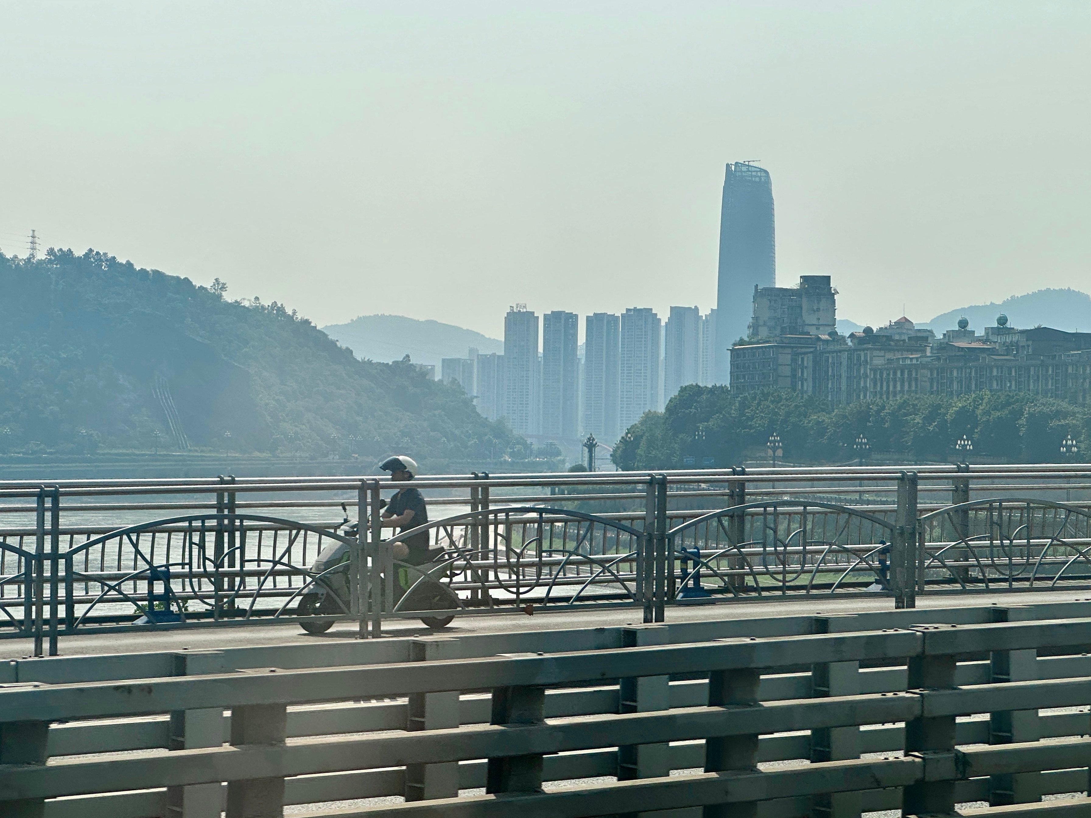 A man rides an electric bike on a bridge that straddles the Yangtze River in Yibin, Sichuan province. Skyscrapers loom against hazy skies in the background. Photo: Frank Chen