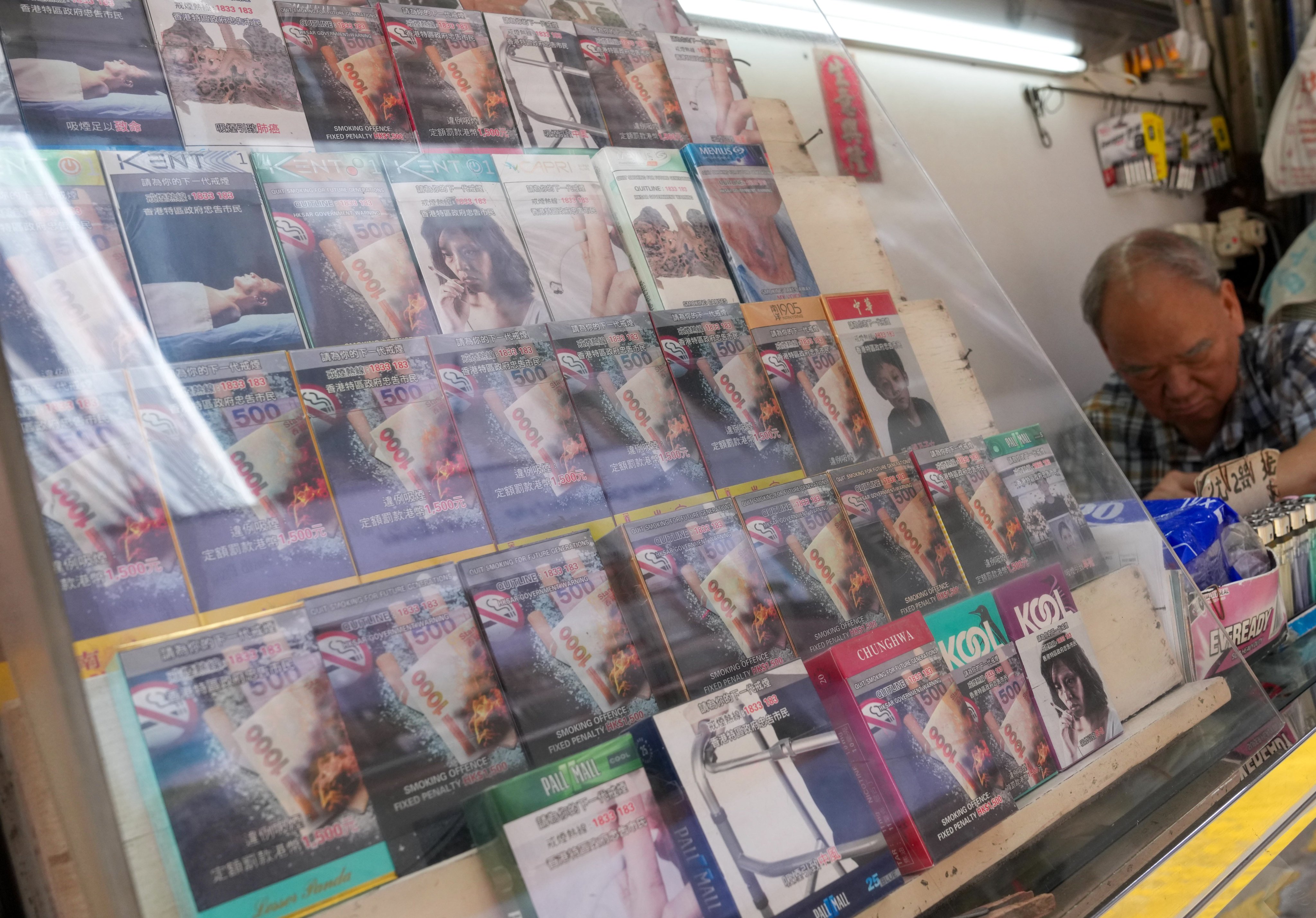 An elderly shopkeeper sits behind a glass display counter packed with cigarette packs featuring graphic health warnings at a traditional newspaper and convenience stall in Hong Kong. Photo: Jelly Tse