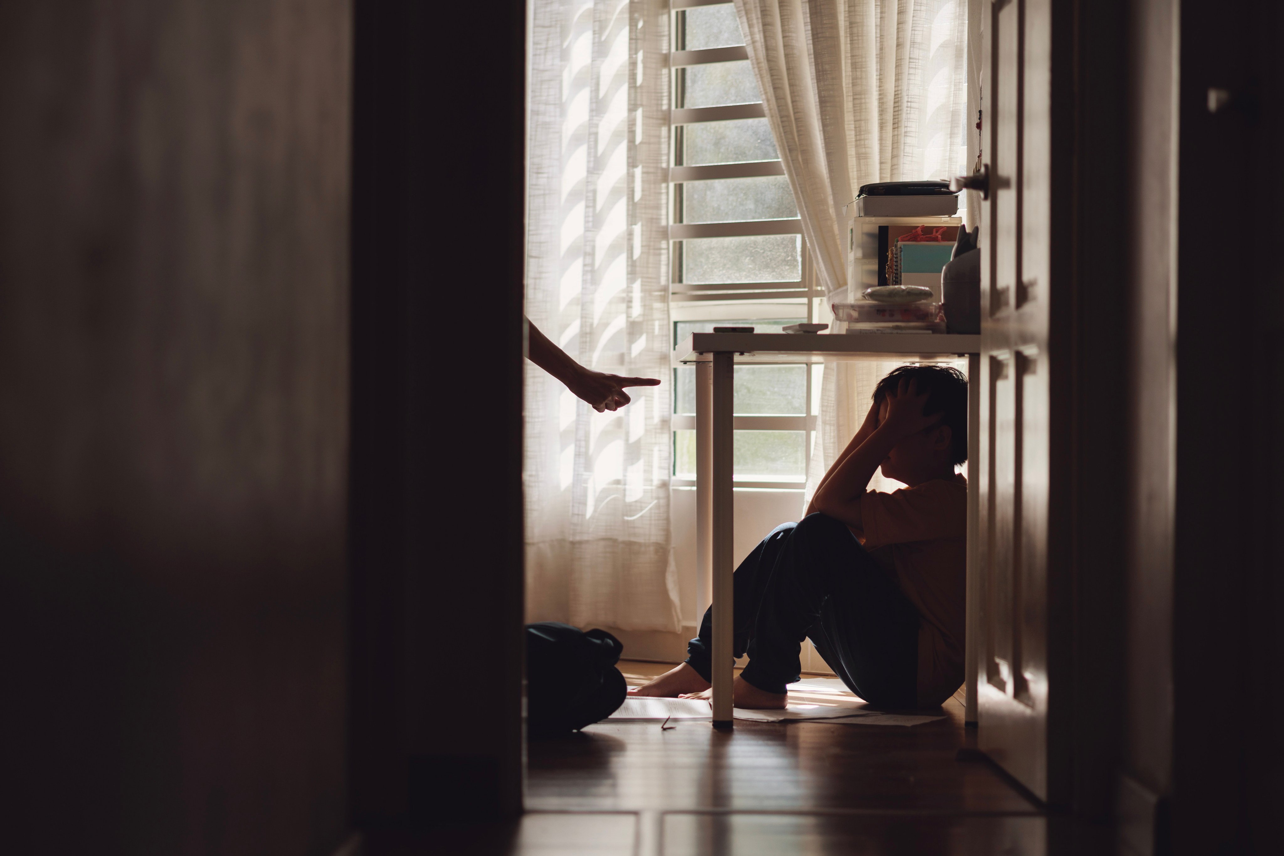 A little Asian boy with a sad and lonely expression hides in the shadows under a table at home, while being scolded by his mother. Photo: Getty Image