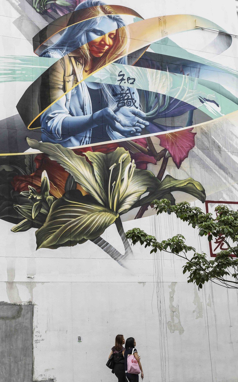 A mural by the artist Fluke on the Hong Kong Institute of Vocational Education, at 6 Oi Kwan Road. Hailing from Montreal, Canada, Fluke excels in large-scale murals that blend photo­realistic subjects with bands of colour and giant brush­strokes. The woman featured here is the artist’s teacher sister, a nod to the piece’s location among schools. The message in her hands reads “knowledge”. Photo: Martin Chan