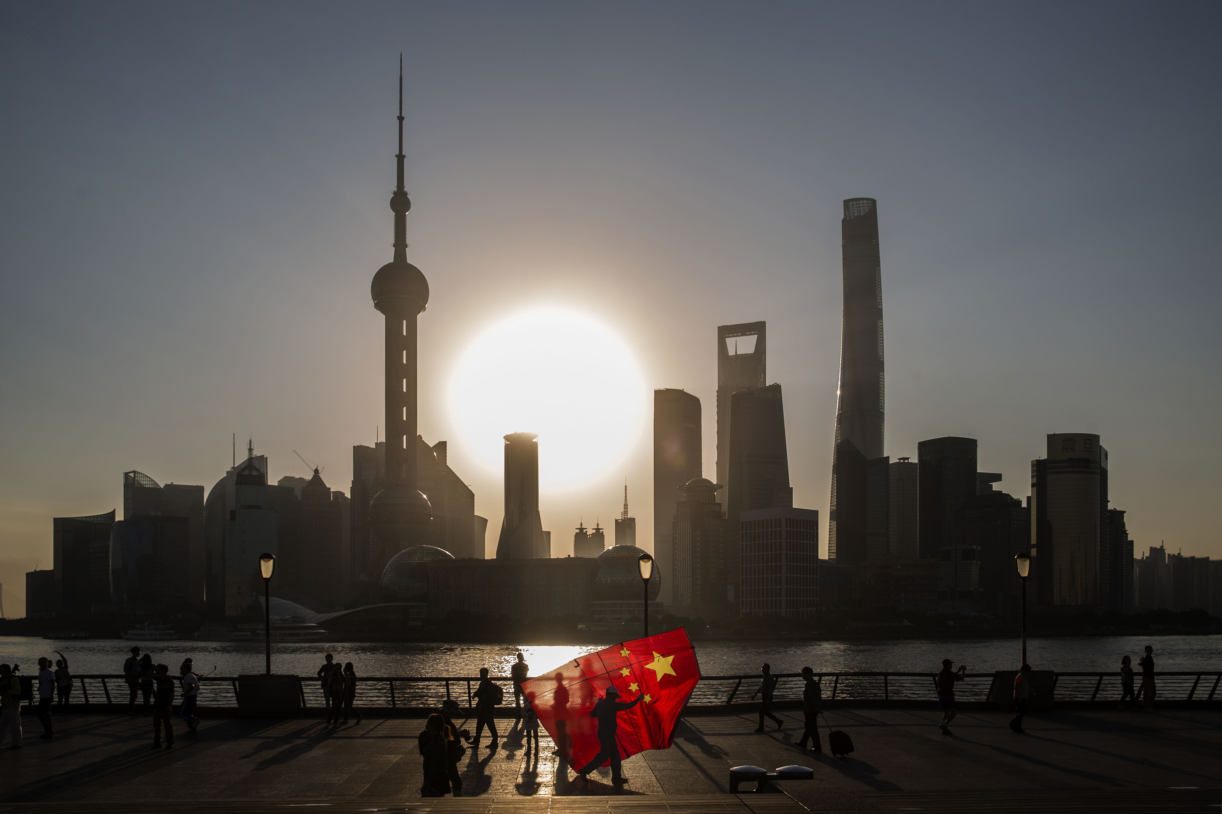 A man carrying a kite in the shape of the Chinese national flag walks along the Bund as the sun rises behind the buildings of Pudong’s Lujiazui financial district in Shanghai in October 2015. China is seeking to build heavyweight investment banks to take on foreign players. Photographer: Bloomberg