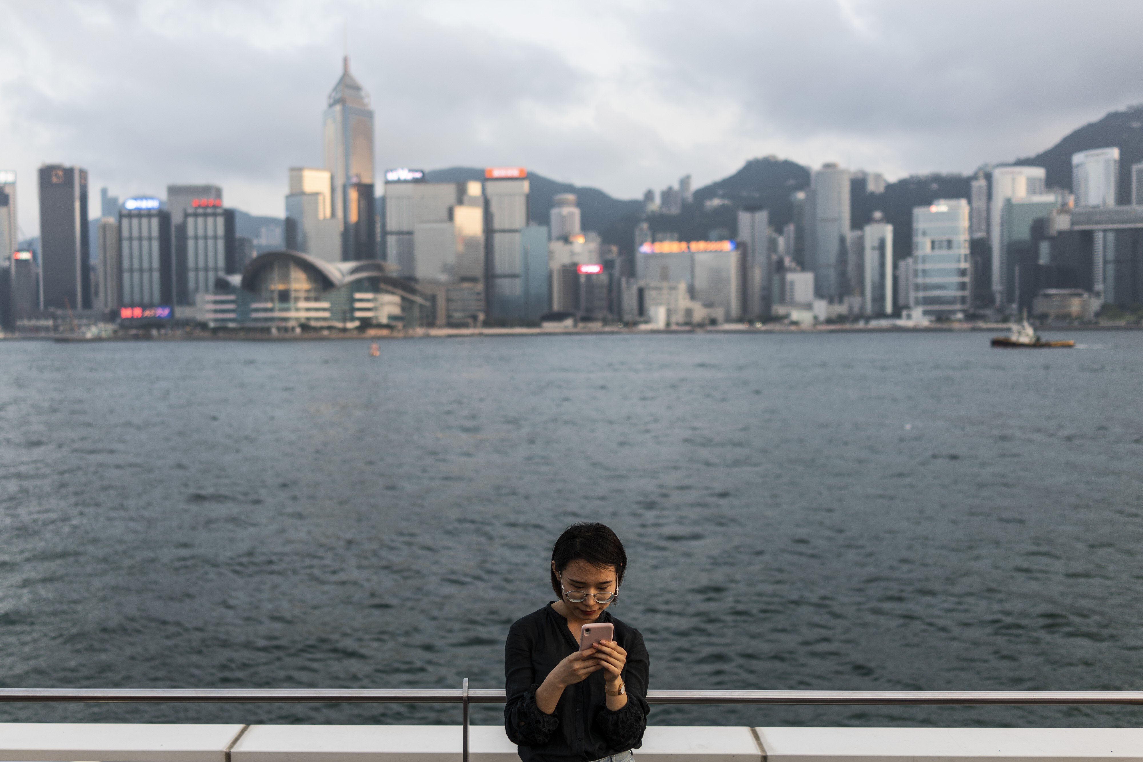 A woman uses a smartphone along Victoria Harbour in Tsim Sha Tsui, flanked by buildings in Hong Kong’s central business district, on April 29, 2019. With the clock ticking on Greater Bay Area integration, Hong Kong is under pressure to upgrade its data governance framework to fit in with China’s digital strategy. Photo: Bloomberg