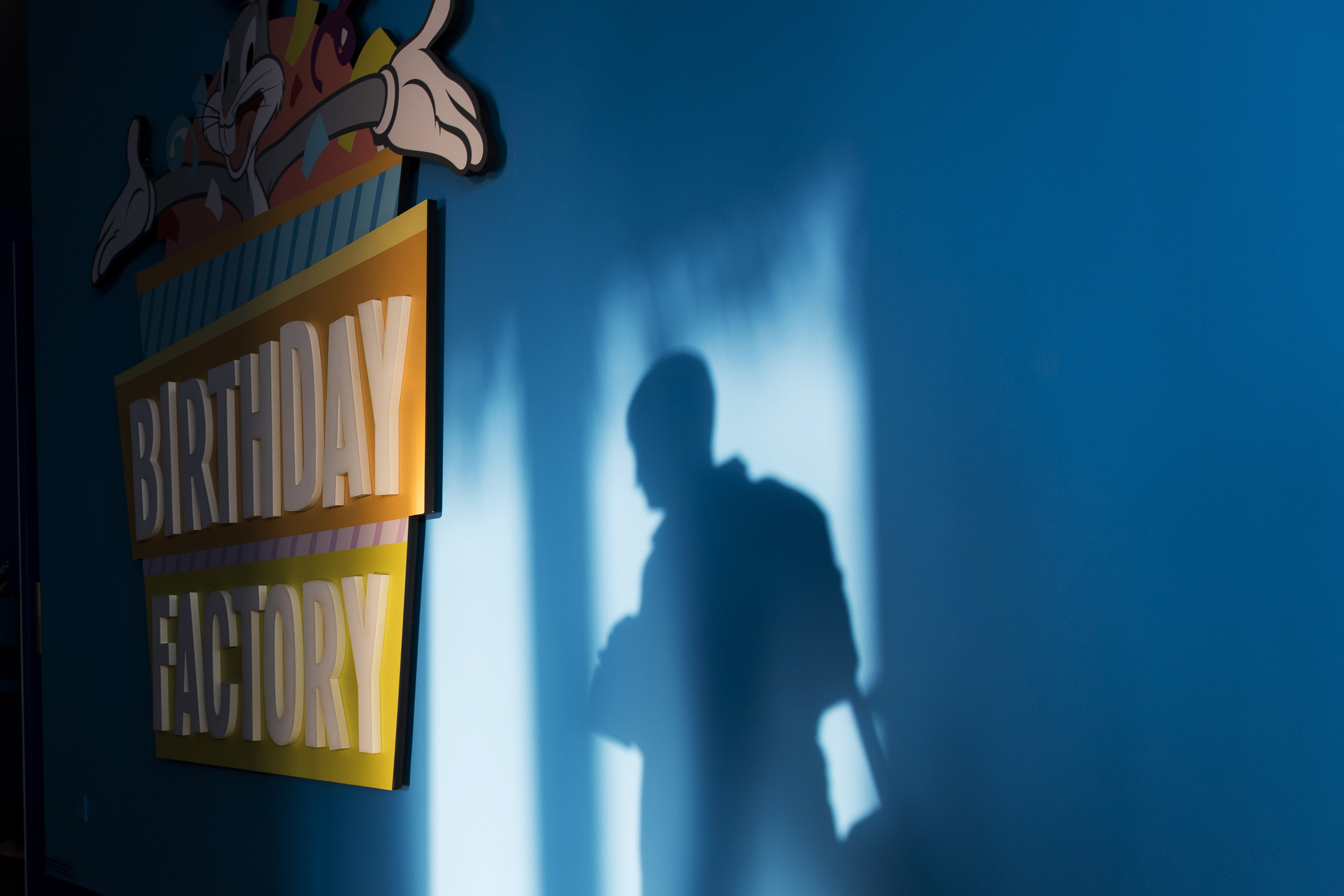 An attendee casts a shadow on a wall next to signage for the birthday Factory in the Warner Bros Fun Zone at the Studio City casino resort, developed by Melco Crown Entertainment Ltd., ahead of the grand opening during a media tour in Macau, China, on Monday, Oct. 26, 2015. Melco Crown is touting a Batman ride, Asia's highest ferris wheel and other Hollywood-centric features for Studio City which is scheduled to open on Oct. 27. Photographer: Xaume Olleros/Bloomberg