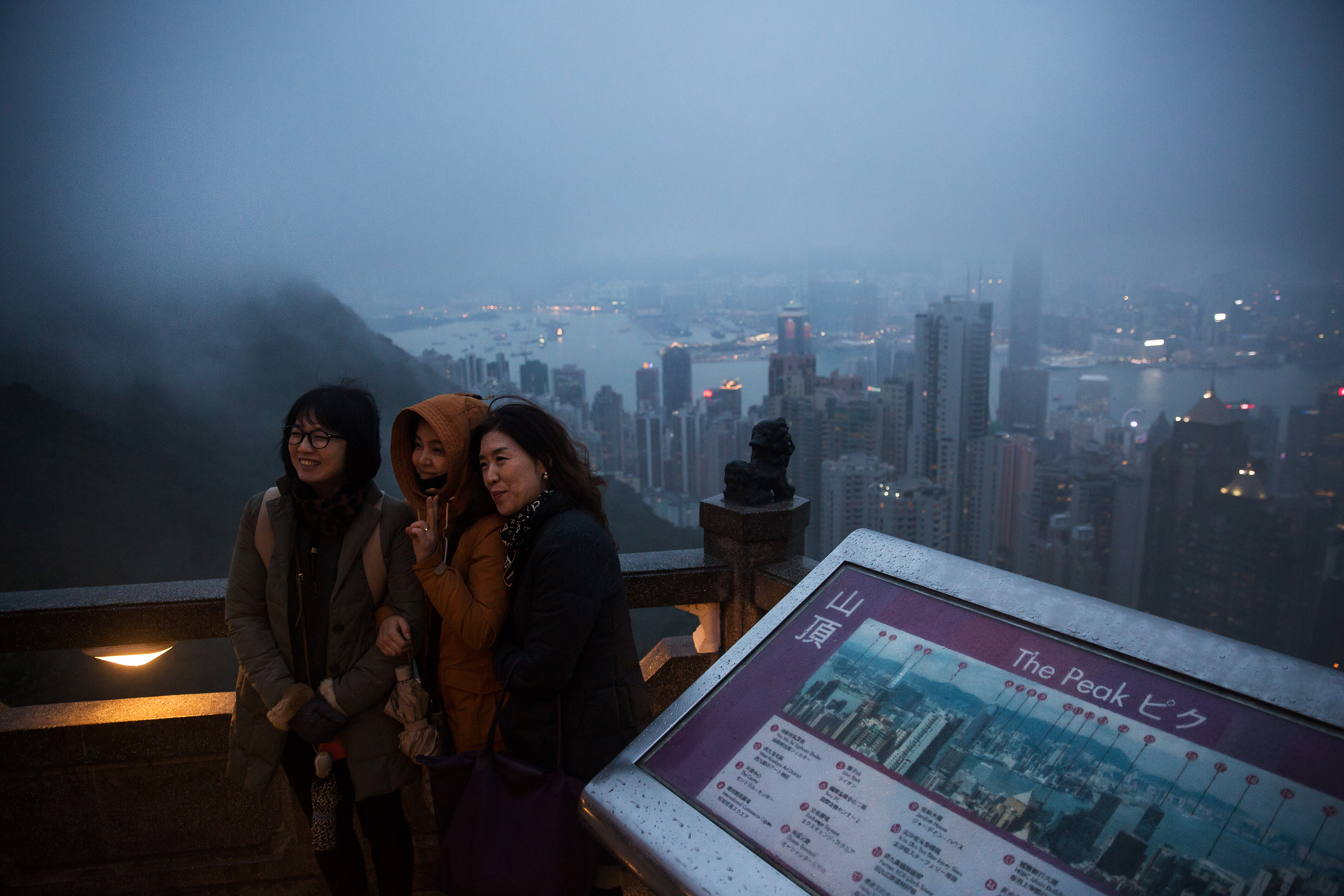 Tourists pose for a photograph as buildings in the background stand shrouded in haze on Victoria Peak in Hong Kong, China, on Friday, Jan. 22, 2016. Hong Kong is expected to grow at a slower pace this year than in 2015 as the city's economy is constrained by difficulties in its exports sector and lower spending by visiting tourists, according to K.C. Chan, Hong Kong's secretary for financial services and the treasury. Photographer: Billy H.C. Kwok/Bloomberg