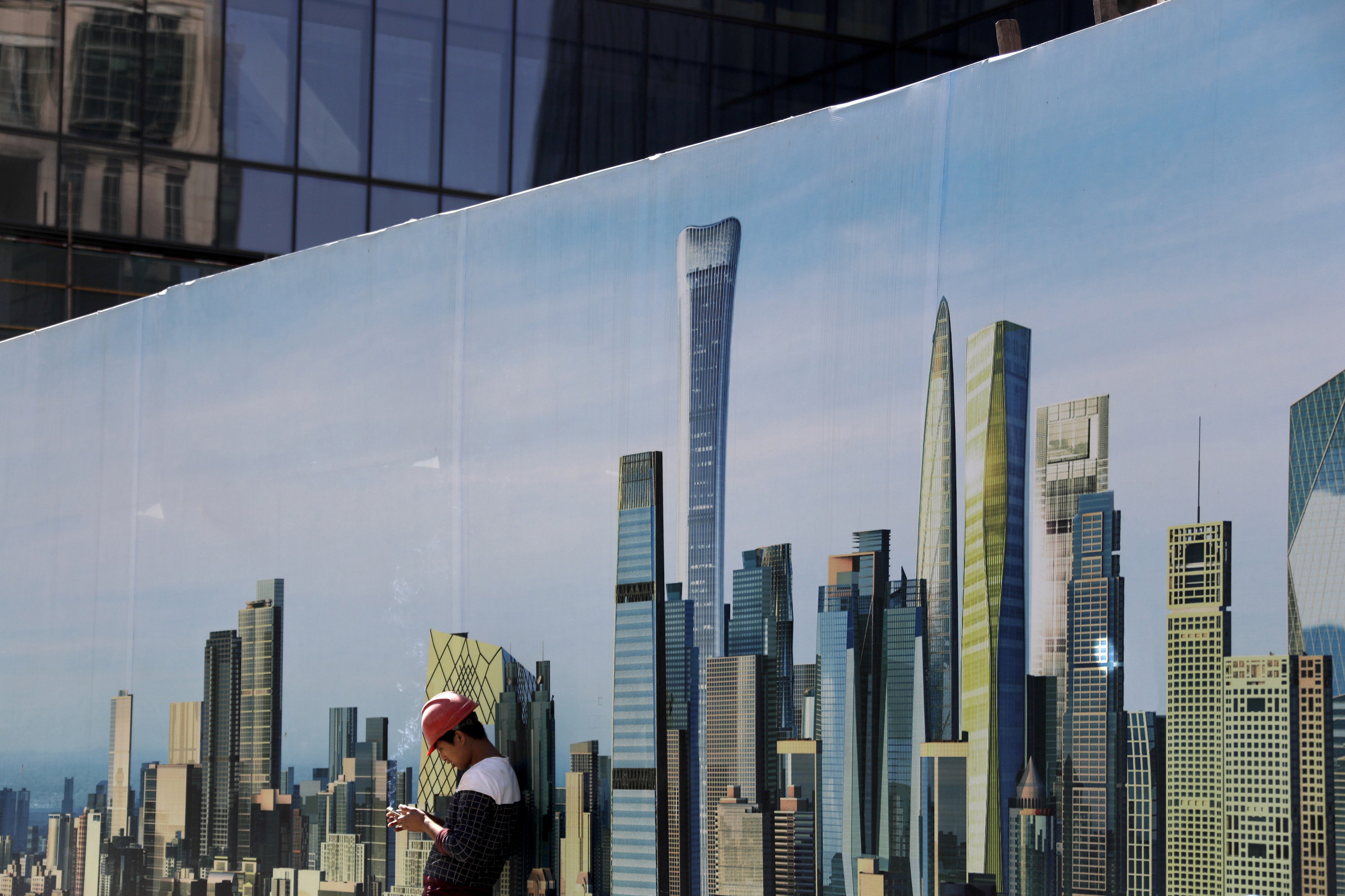 A worker smokes while checking on his mobile phone at a construction site wall depicting the skyscrapers in the Chinese capital at the Central Business District in Beijing, Tuesday, Sept. 19, 2017. Photo: AP