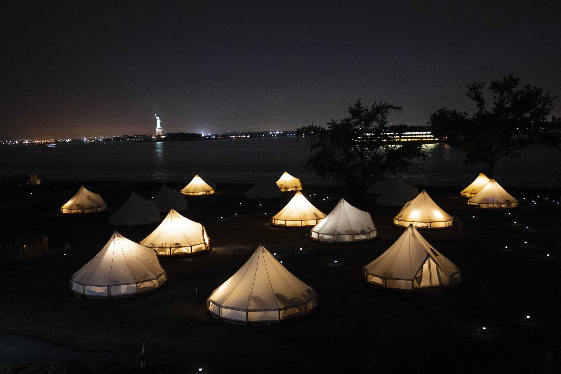 Glowing Journey Tents at night in the foreground with the Statue of Liberty and Staten Island Ferry behind. Photo: David Gray/Bloomberg