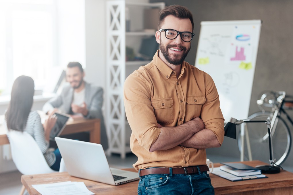 Confident team leader. Confident young man keeping arms crossed and looking at camera with smile while his colleagues working in the background