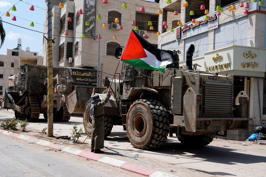 A Palestinian man holds a national flag in front of Israeli military vehicles during an Israeli raid in the Tulkarm refugee camp in the northern West Bank, on Thursday, August 22, 2024. Three Palestinians were killed on Thursday in an Israeli bombing on a house in the Tulkarm refugee camp, according to the Palestinian official news agency WAFA. Photo: Xinhua
