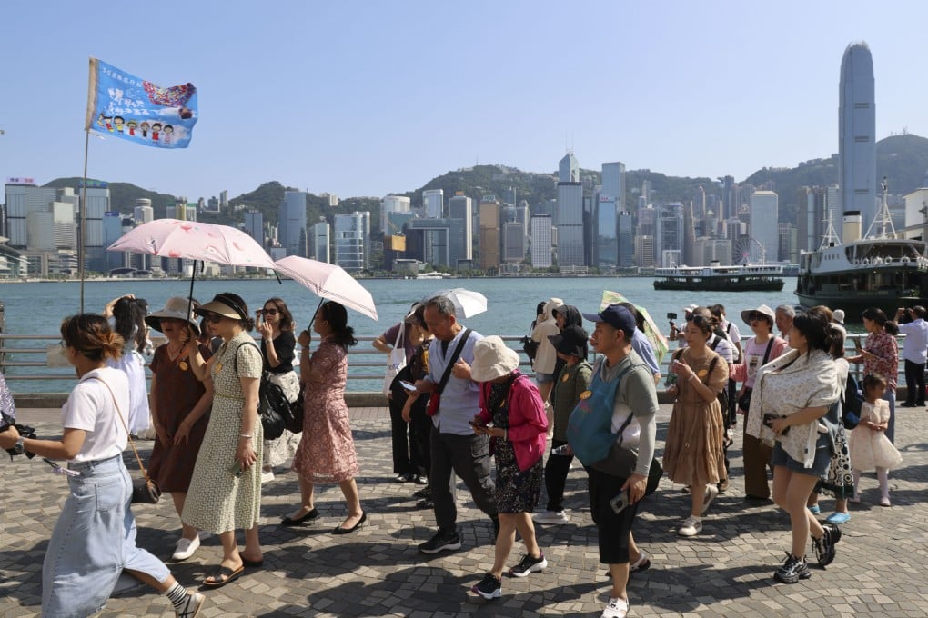 A large group of tourists, including elderly and young people, walk along Victoria Harbour with Hong Kong's skyline in the background, some holding umbrellas to shield from the sun, after a tsunami threat following the Japan earthquake on December 9, 2025.
