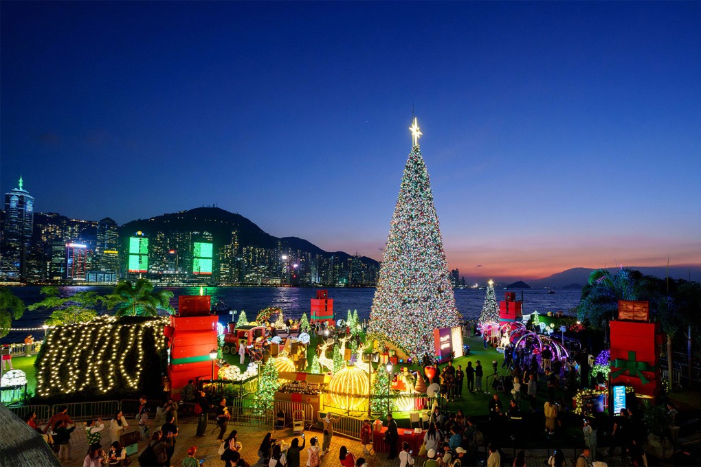 A large, illuminated Christmas tree decorated with colorful lights and topped with a star is surrounded by festive LED light displays, gifts, and crowds of people at a holiday market against a backdrop of Hong Kong's skyline at dusk.