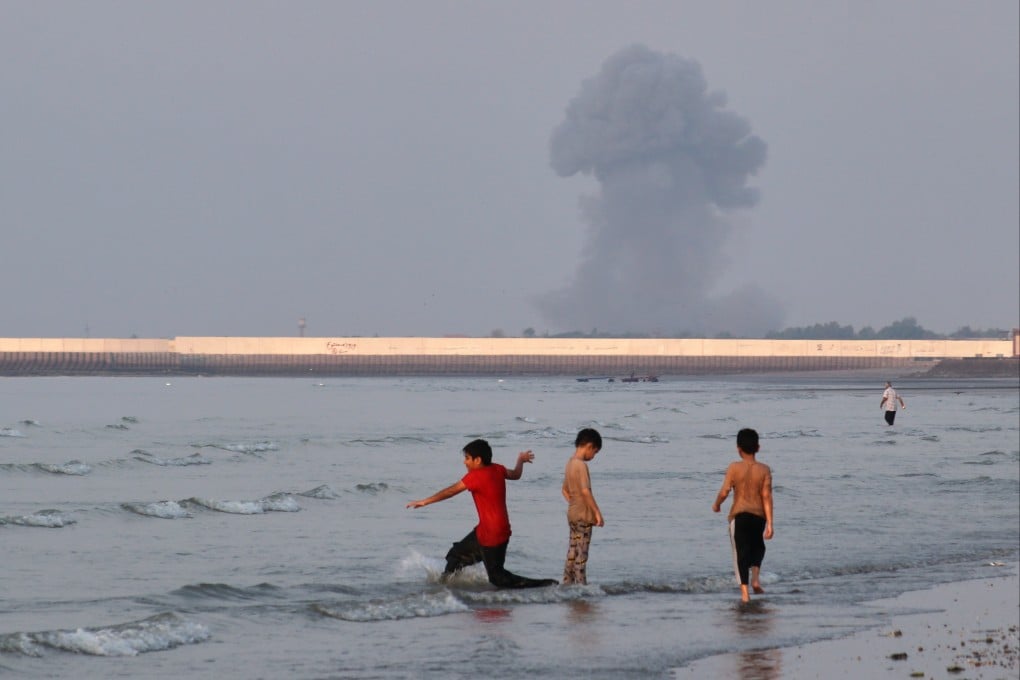 Boys play in the shallow waters of the Strait of Hormuz, as a plume of smoke rises from an explosion in the background, off Bandar Abbas, Iran, on Monday. Photo: ISNA via AP