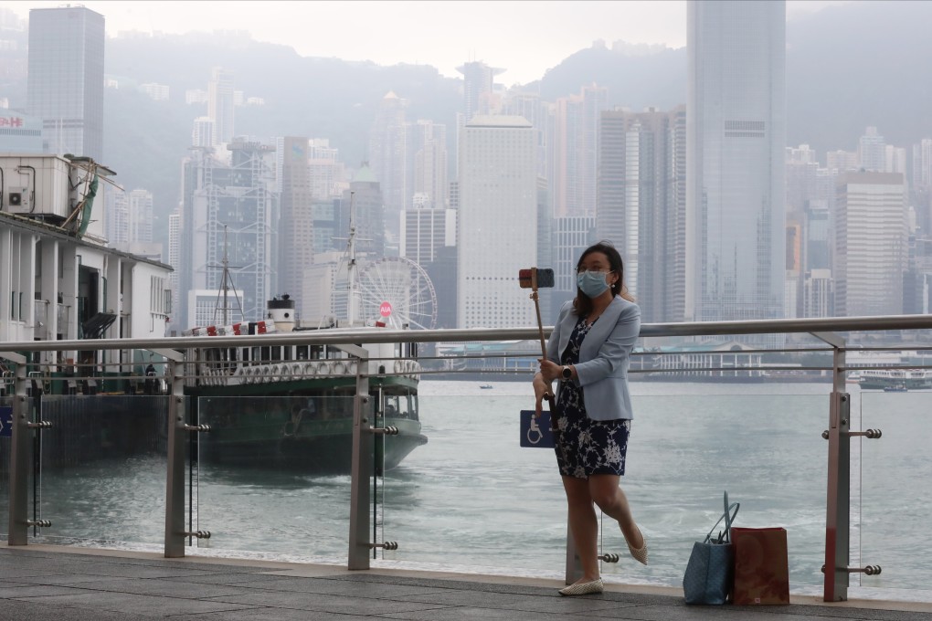 A woman in a face mask with a selfie stick poses for a photo with the Victoria Harbour and Hong Kong skyline in the background. Photo: K.Y. Cheng