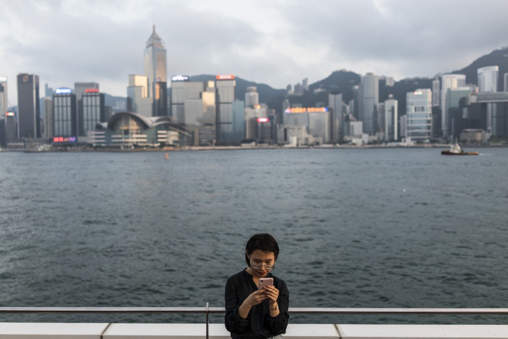A woman uses a smartphone along Victoria Harbour in Tsim Sha Tsui, flanked by buildings in Hong Kong’s central business district, on April 29, 2019. With the clock ticking on Greater Bay Area integration, Hong Kong is under pressure to upgrade its data governance framework to fit in with China’s digital strategy. Photo: Bloomberg
