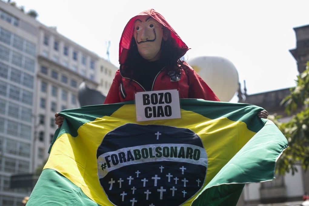 A demonstrator, wearing a Money Heist mask and costume, holds a Brazilian flag with a message that reads in Portuguese “Get out Bolsonaro” during a protest in Rio de Janeiro, Brazil on Saturday. Photo: AP