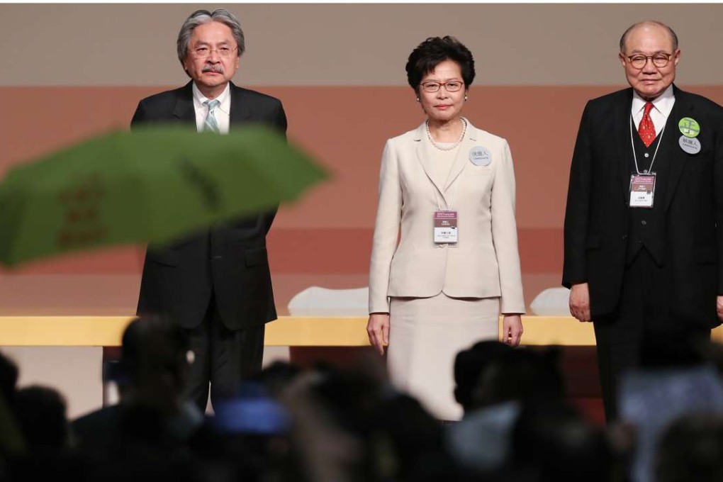 A yellow umbrella is raised in defiance as Carrie Lam stands on stage with election rivals John Tsang and Woo Kwok-hing. Photo: Robert Ng