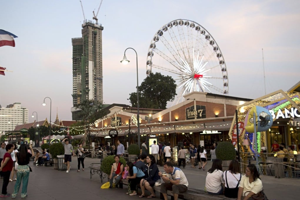 Visitors sit on a bench in front of restaurants as a ferris wheel operates in the background at Asiatique The Riverfront open-air mall in Bangkok, Thailand. Photo: Brent Lewin/Bloomberg