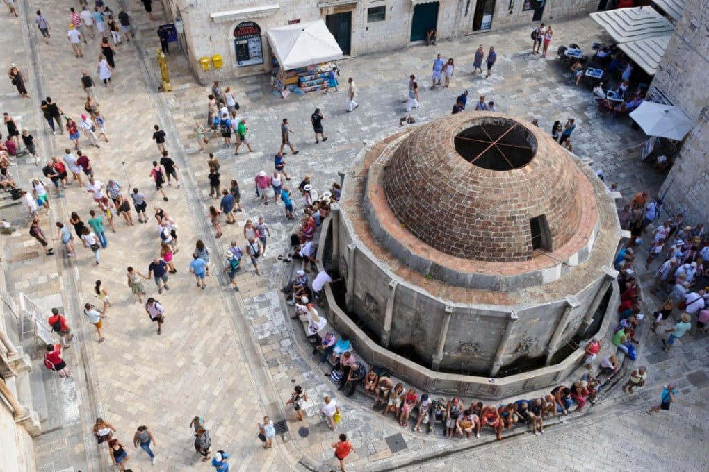 A crowd of tourists gather around the Onofrio fountain at the entrance of the fortress in the Old Town, Dubrovnik. The city is being overrun by tourists. Photo: Alamy