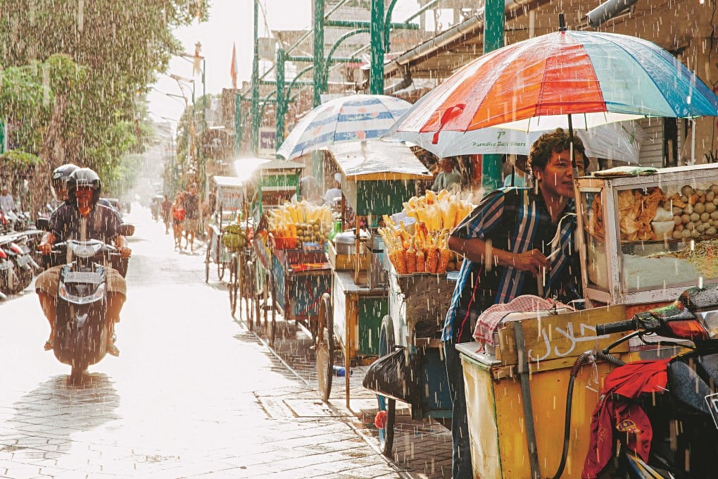 A food vendor hides from the heavy rain under his stall’s umbrella on a street in Kuta, Indonesia. The holiday island saw unseasonal rains this year. Photo: Alamy