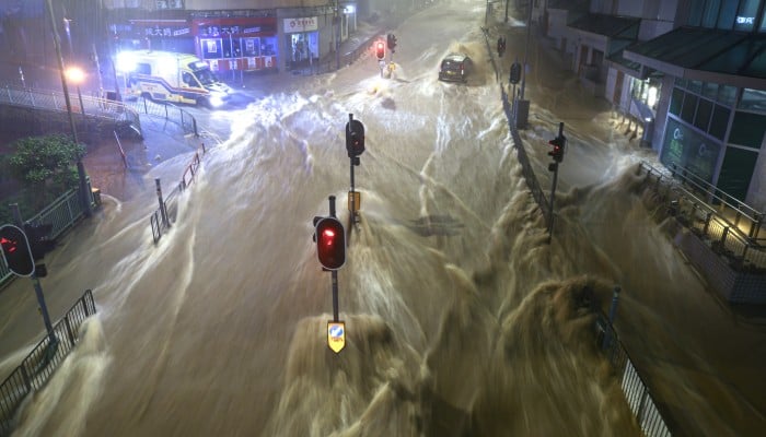 Floodwaters from heavy rain during a Black Rainstorm Warning Signal surge through an urban intersection in Hong Kong, submerging traffic lights, cars, and sidewalks.