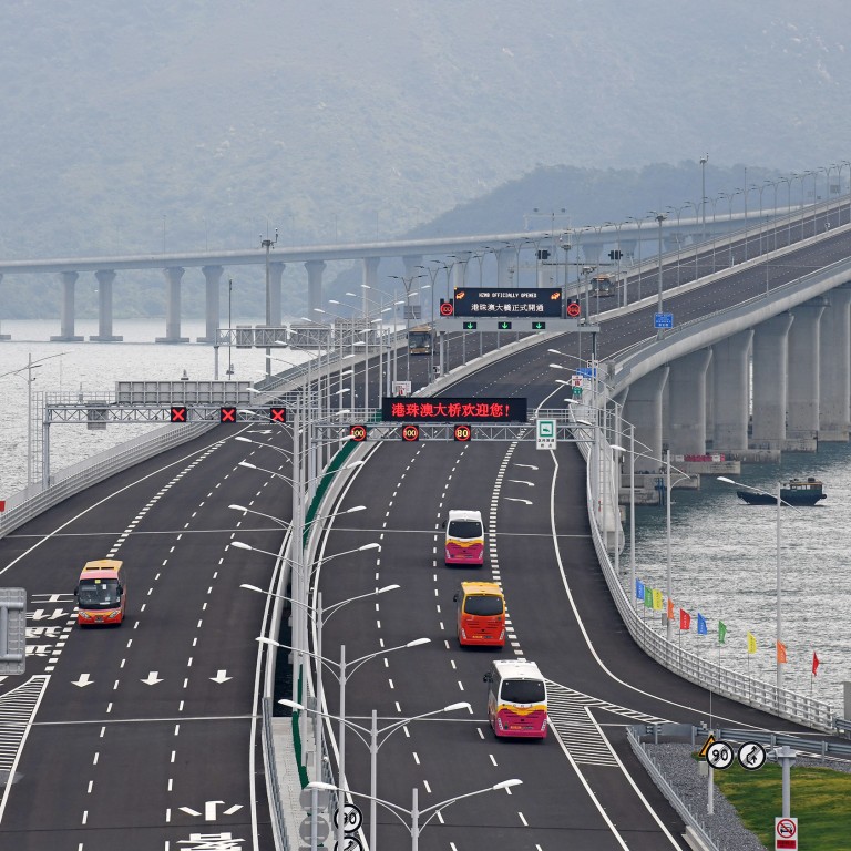 A series of buses and vehicles travel on a newly constructed road near the water, with traffic signals and signs indicating cross-border driving regulations in Hong Kong.