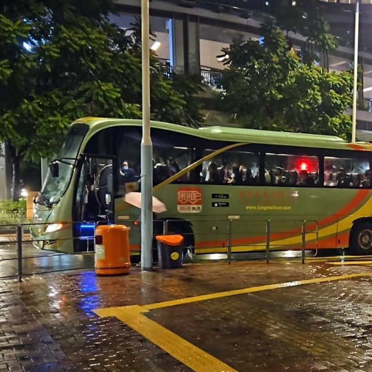 A green double-decker bus with passengers onboard is stopped at a bus stop in rainy weather, with wet pavement, trash bins, a street lamp, and lush trees in the background.
