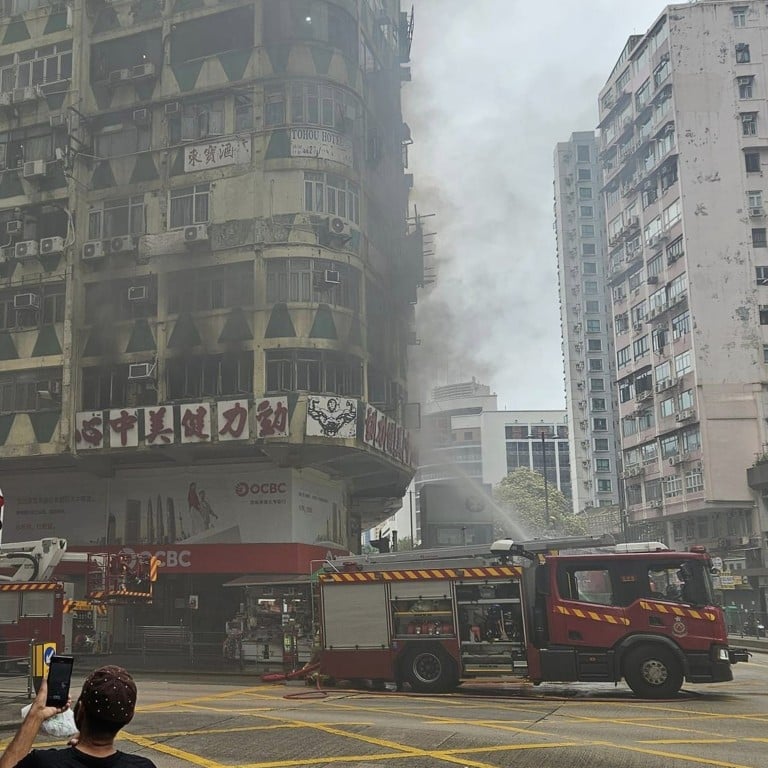 A fire truck is spraying water onto a burning, smoke-filled building in a densely populated city area, with bystanders observing nearby.