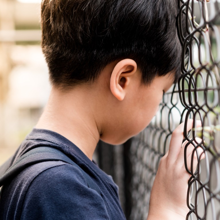 A young boy with short dark hair is standing close to a chain-link fence, with his head bowed and hand resting on the fence, appearing somber or contemplative.
