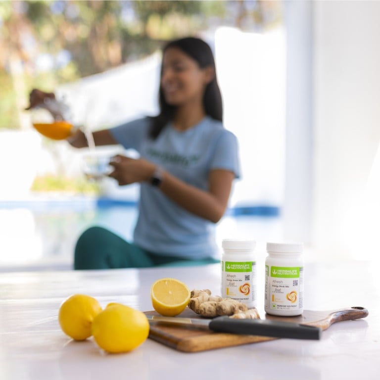 Woman smiling, pouring juice, with lemon, ginger, and supplements on a table in a bright kitchen.