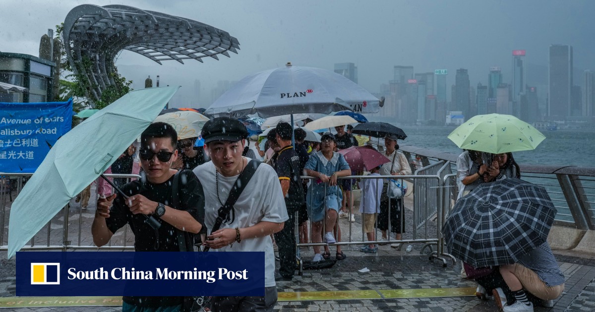 A crowd of people, many holding umbrellas, gathers along a waterfront with a backdrop of skyscrapers under heavy rain and a cloudy sky, reflecting the impact of Super Typhoon Ragasa at Hong Kong International Airport.