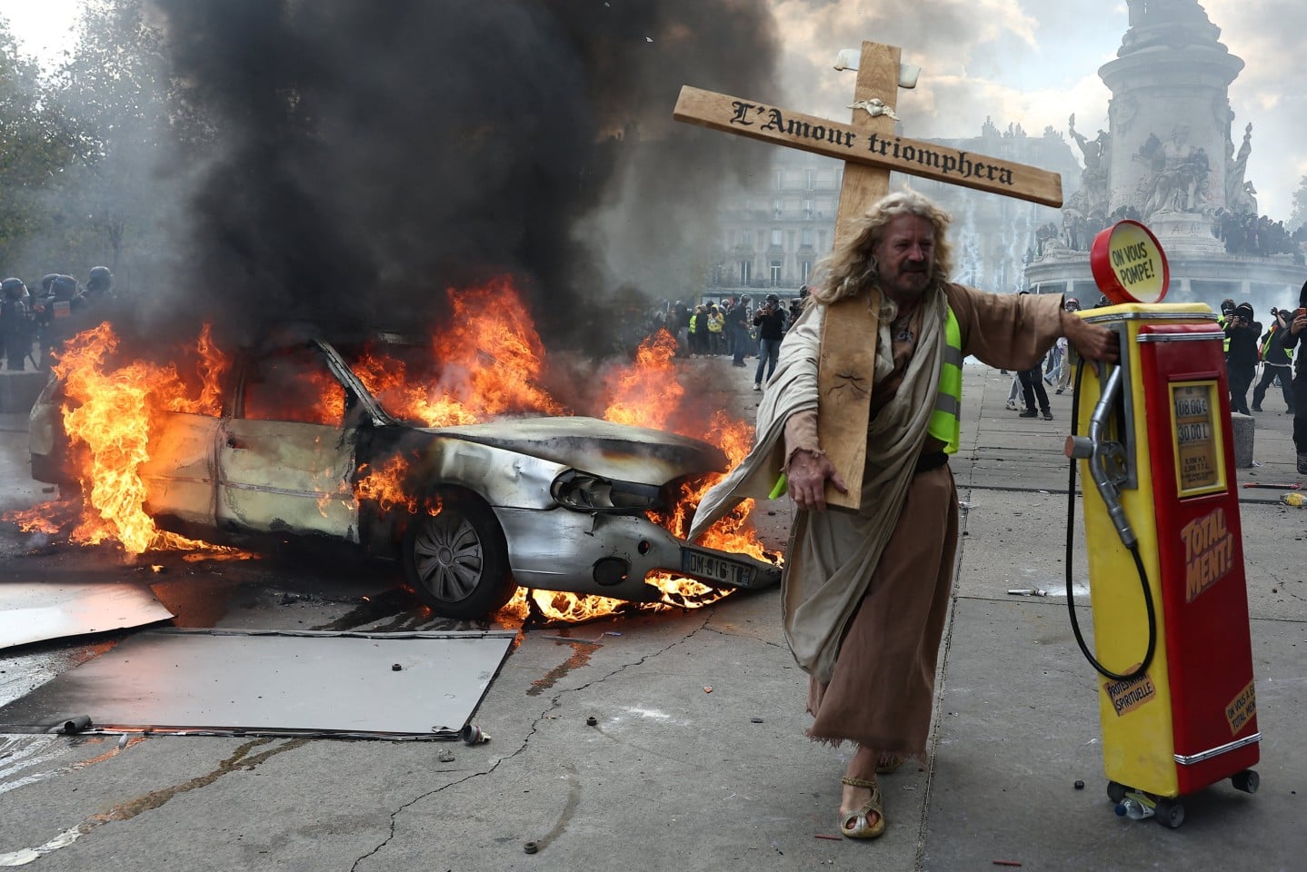 A protester dressed as Jesus Christ and holding a mock gas station dispenser walks past a burning car. Photo: Reuters