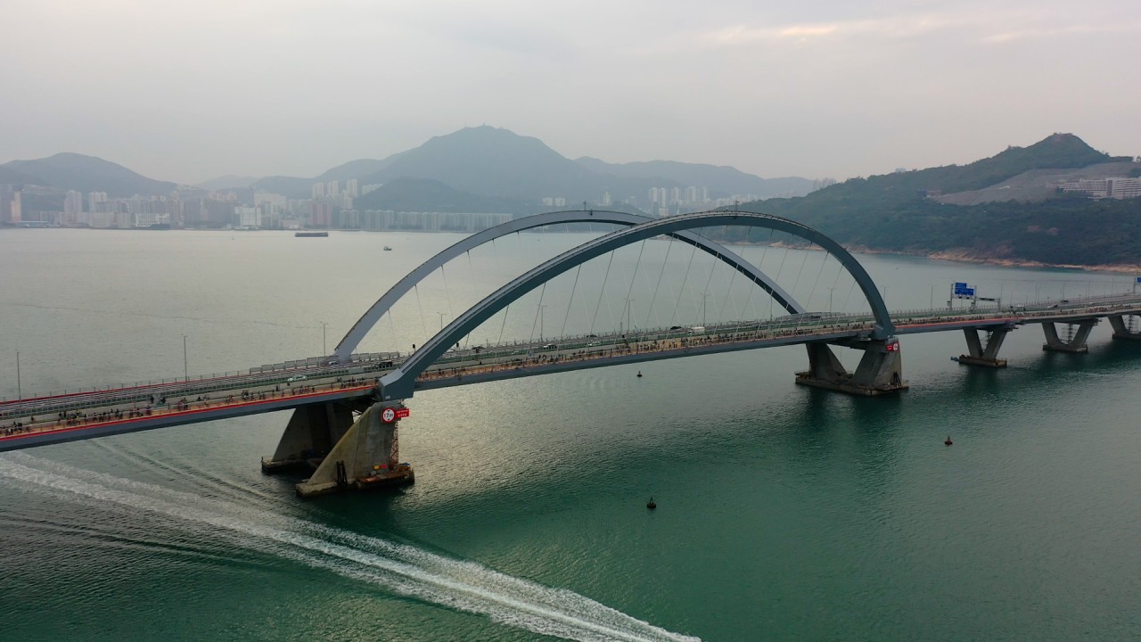 The image shows a modern cable-stayed bridge spanning over a body of water, with mountains and a city skyline in the background under a cloudy sky.